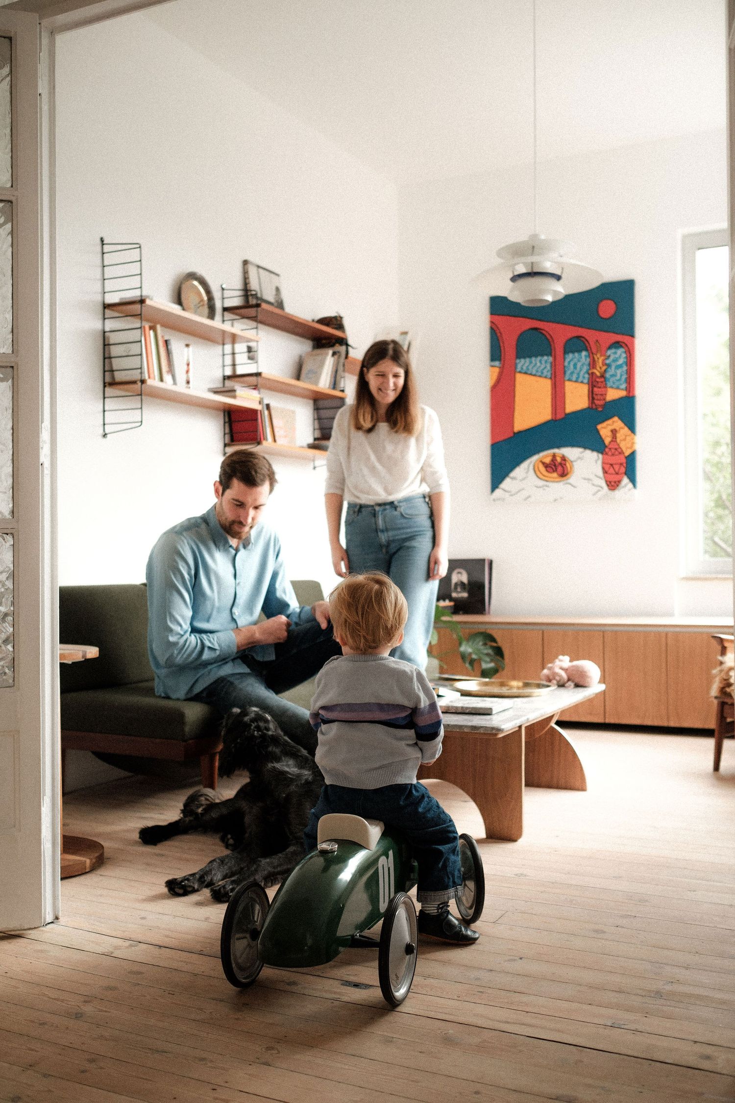 A bright living room scene with a child on a toy vehicle and a black dog resting near modern furniture.