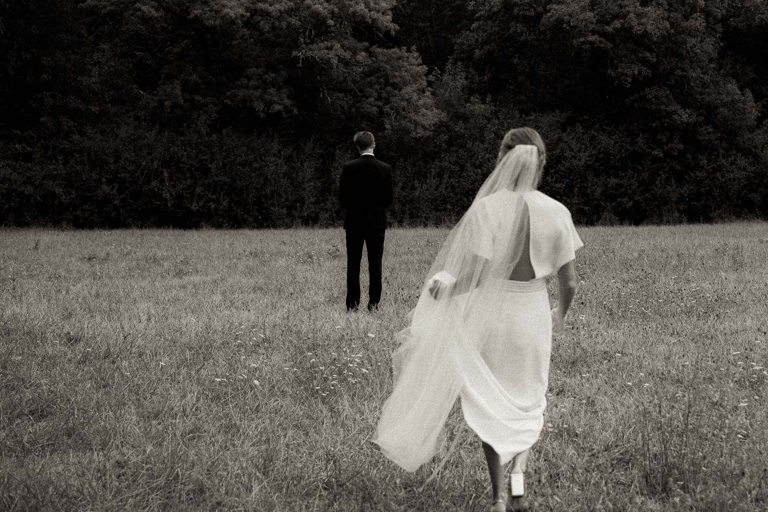 Black and white photo of a bride in flowing dress walking through a meadow towards someone in dark attire.