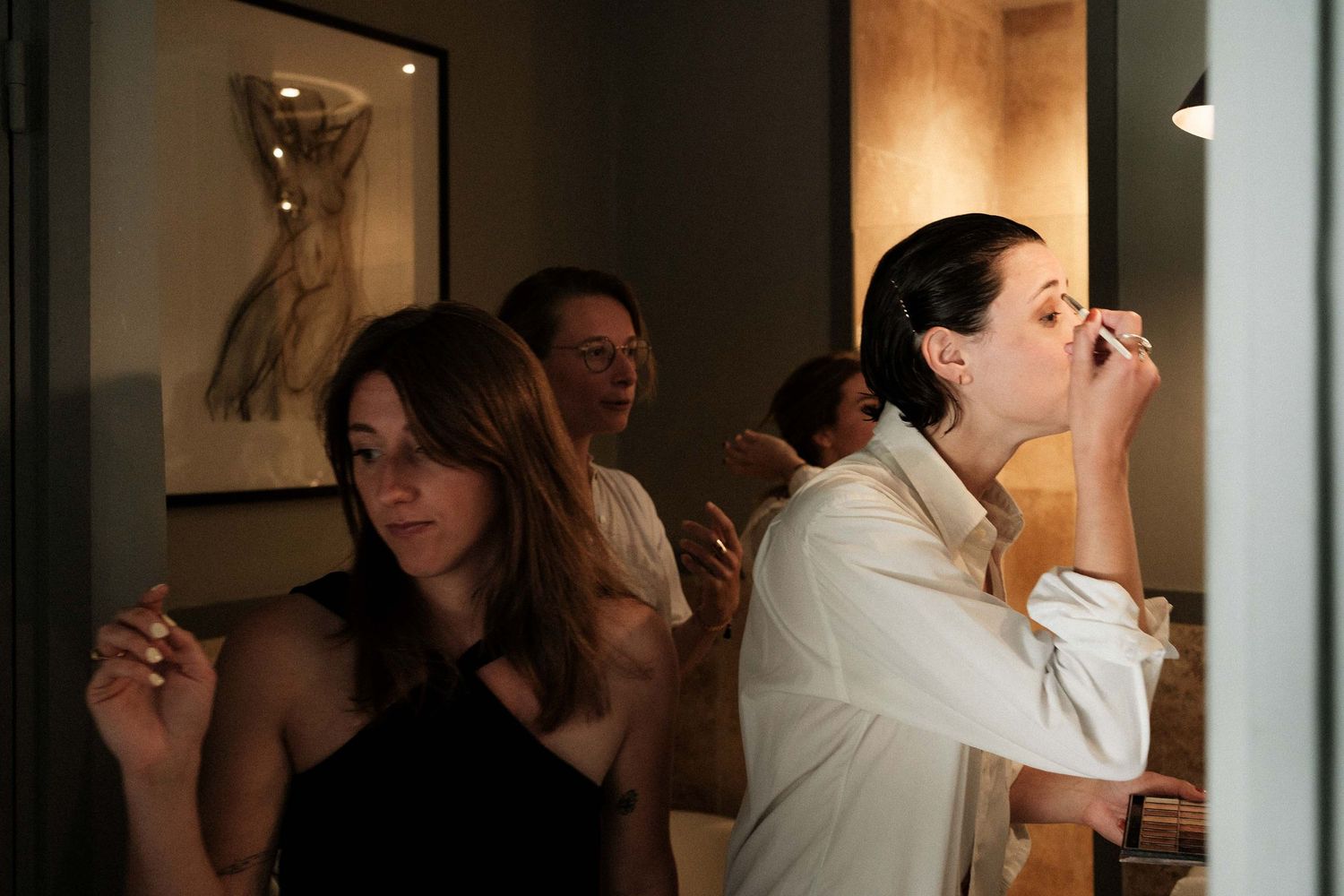A person in a white dress stands in front of a mirror while others help prepare for a wedding ceremony.