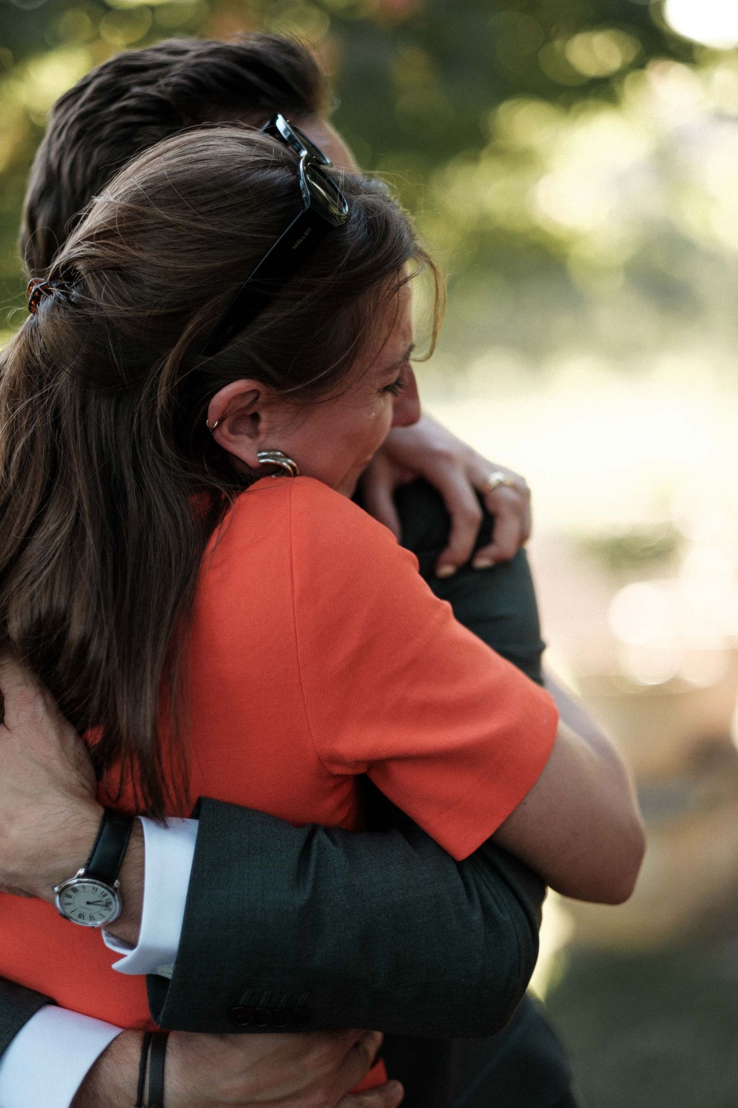 Two people share an emotional embrace outdoors with blurred natural background in warm lighting.