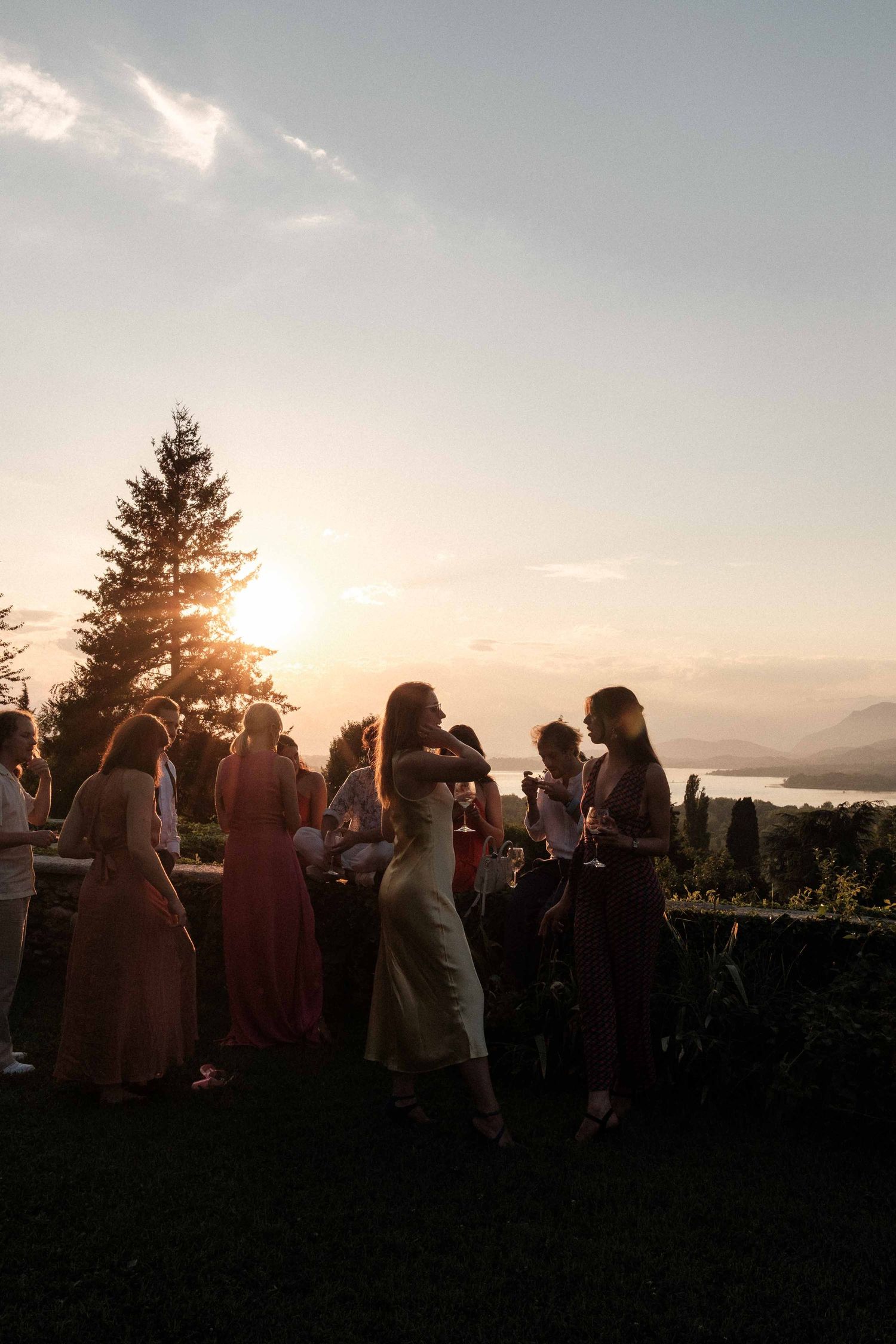A group of people gathered outdoors at sunset with evergreen trees silhouetted against the glowing sky.
