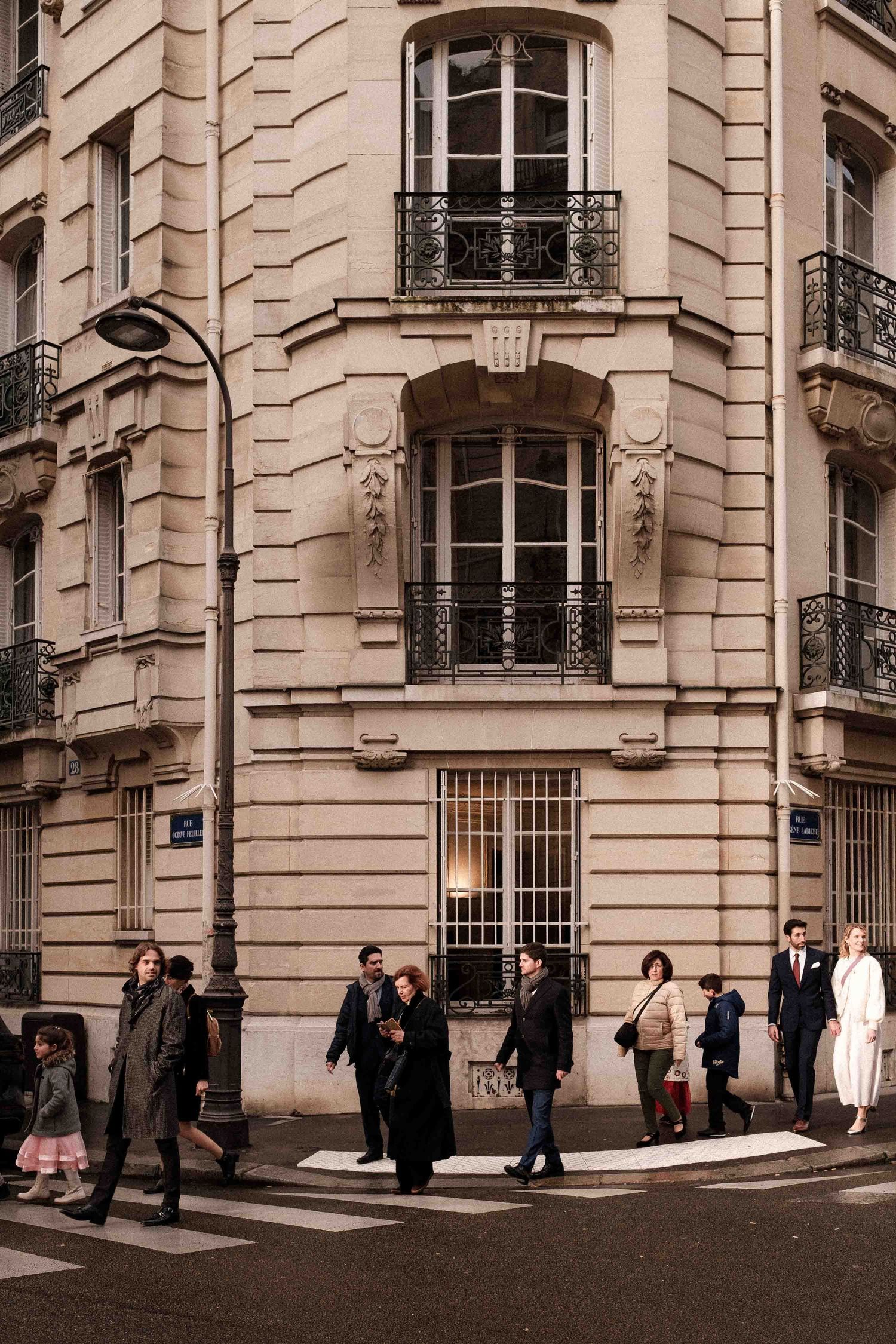 People walking past a classic Parisian corner building with ornate architecture and wrought iron balconies.