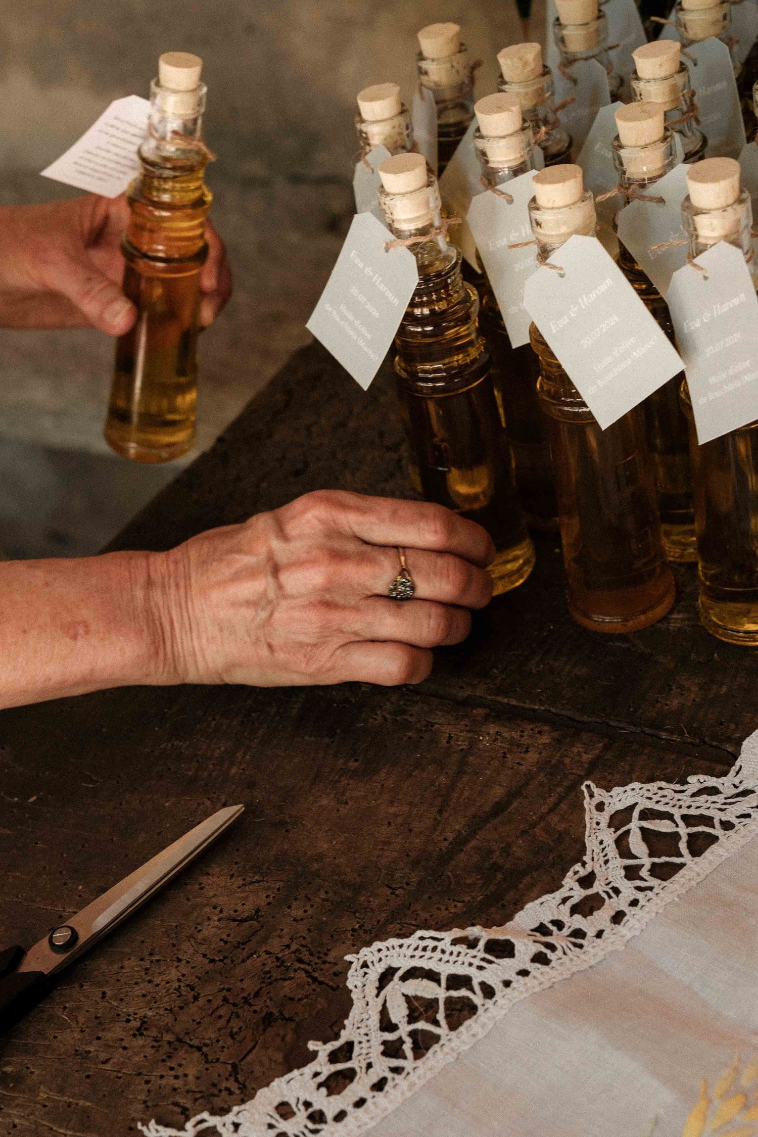 Small glass bottles with cork stoppers and paper tags sit on a dark wooden surface with white lace doily details.