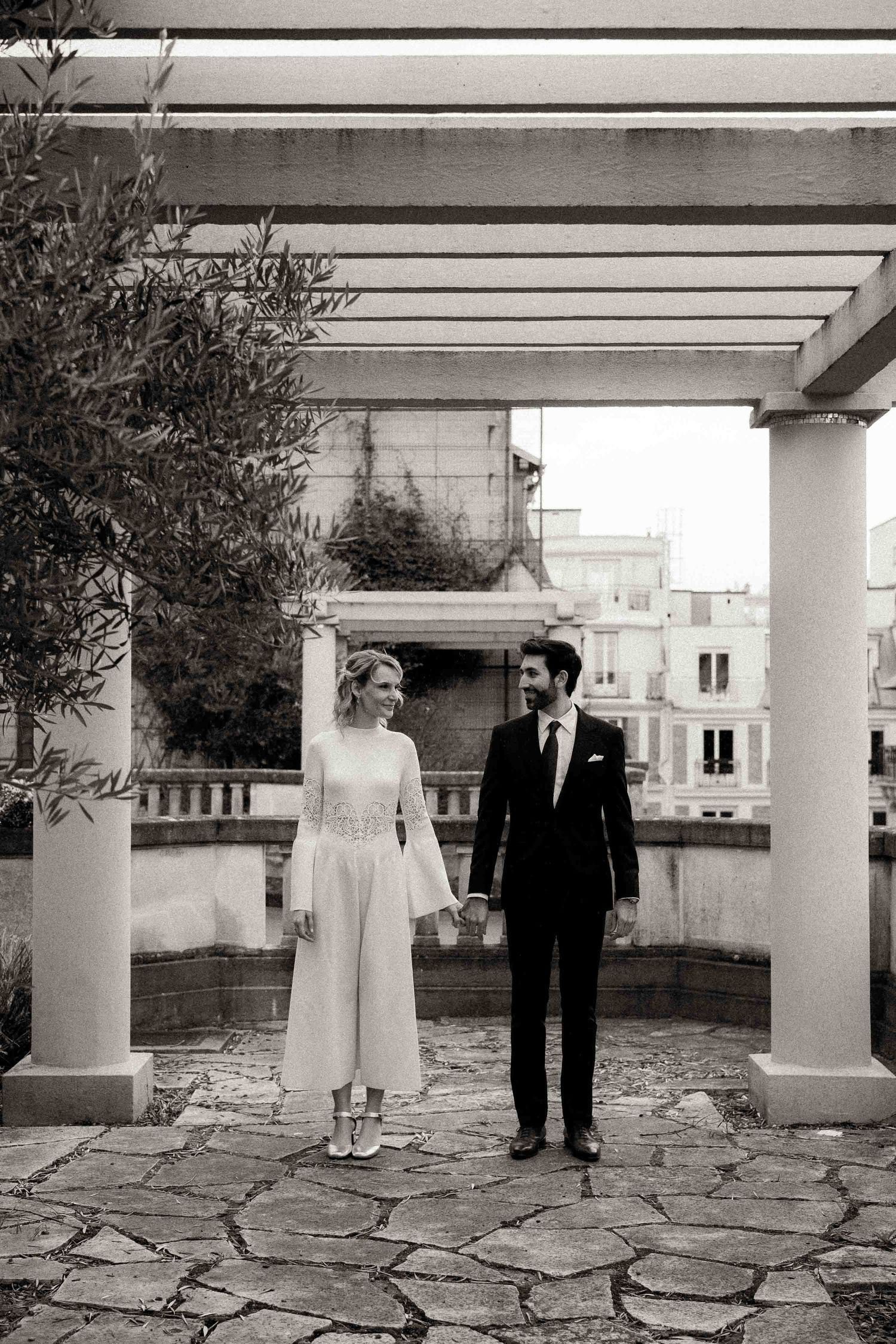 A couple stands together on a stone patio with white columns and railings in a black and white vintage photograph.