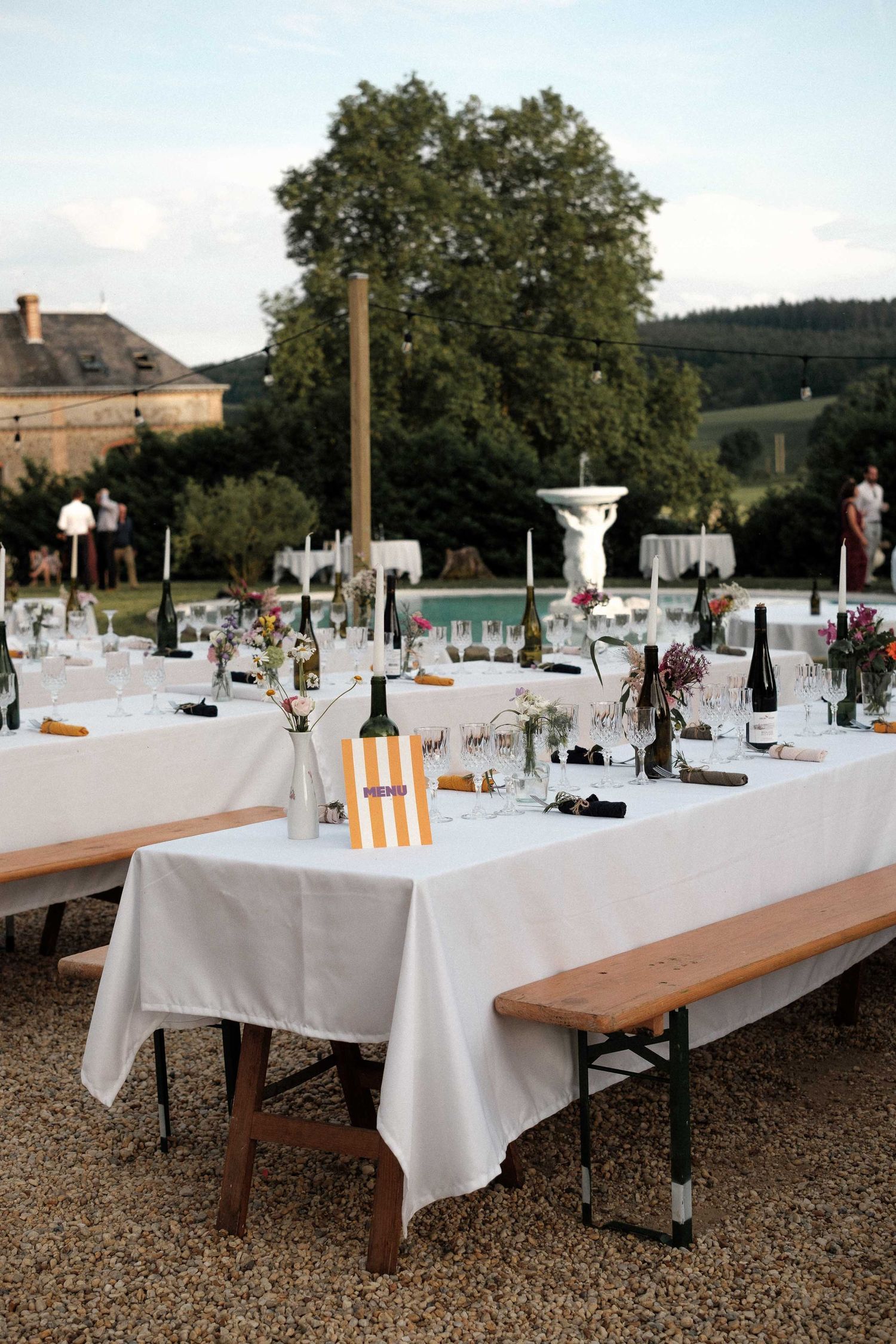 Long rustic wooden benches with white tablecloths set up for outdoor dining at a countryside venue.