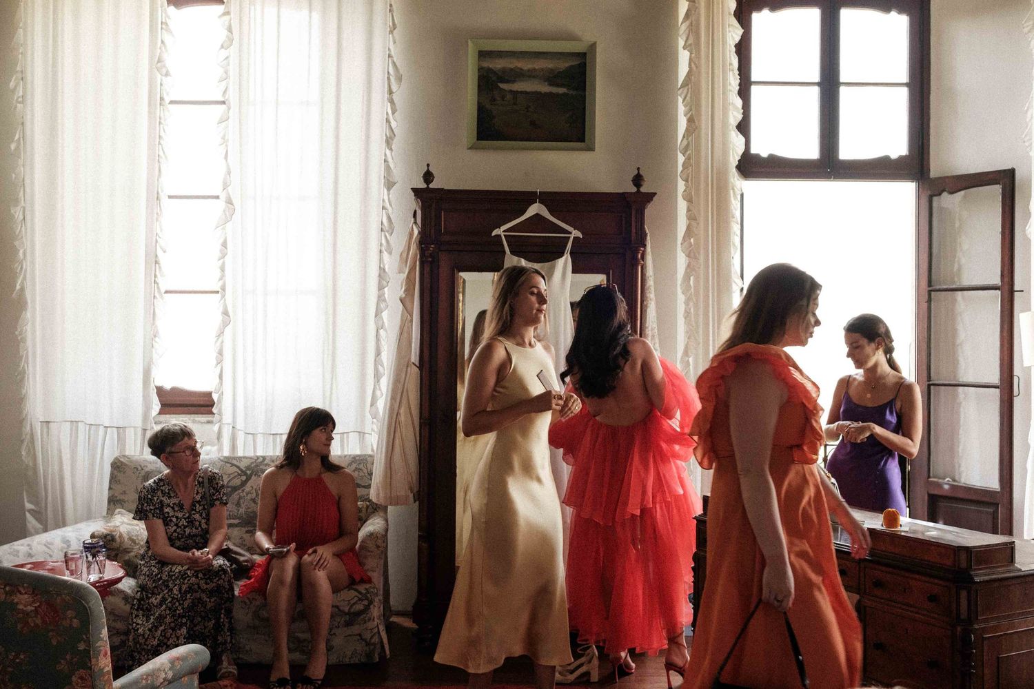 A group of people in formal attire gather near windows in an elegant room with natural lighting.