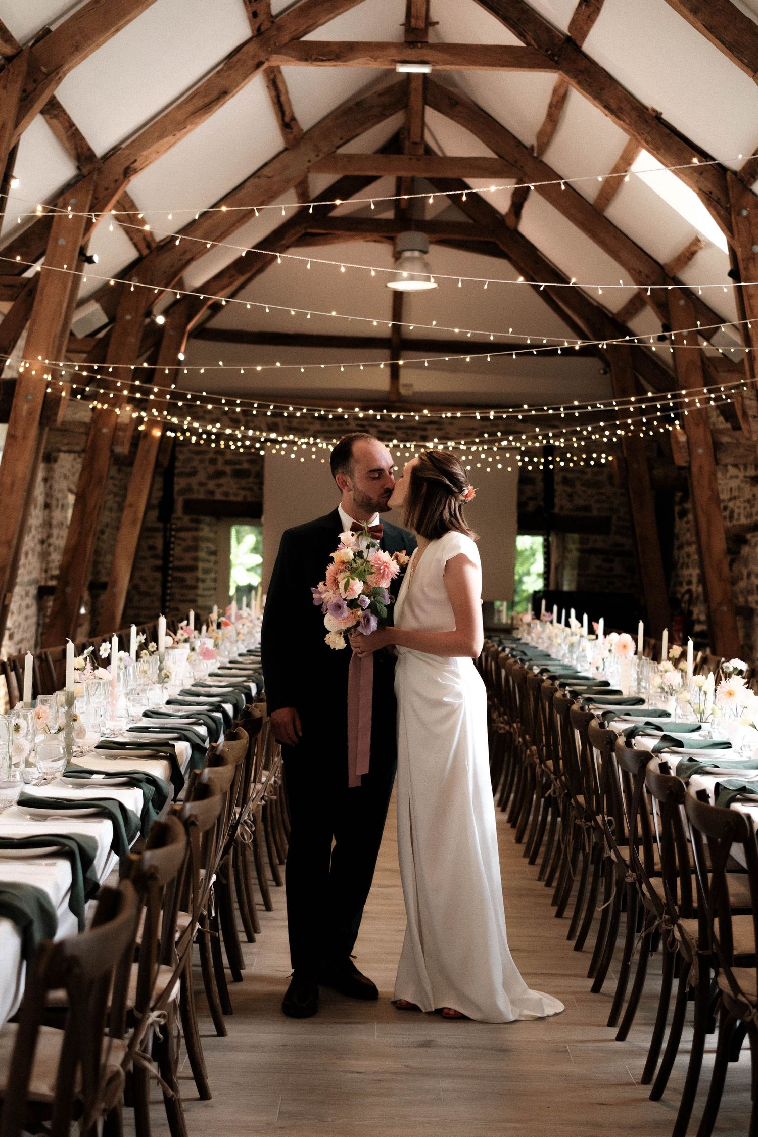 Rustic barn wedding reception venue decorated with string lights and long rows of wooden tables and chairs.