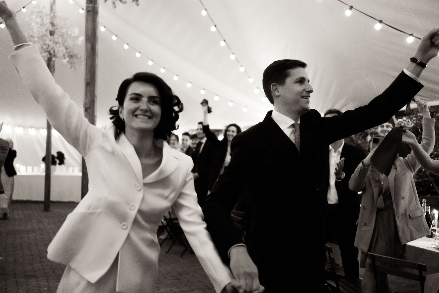 A joyful couple celebrates with raised arms while dancing at their wedding reception under a tent with string lights.