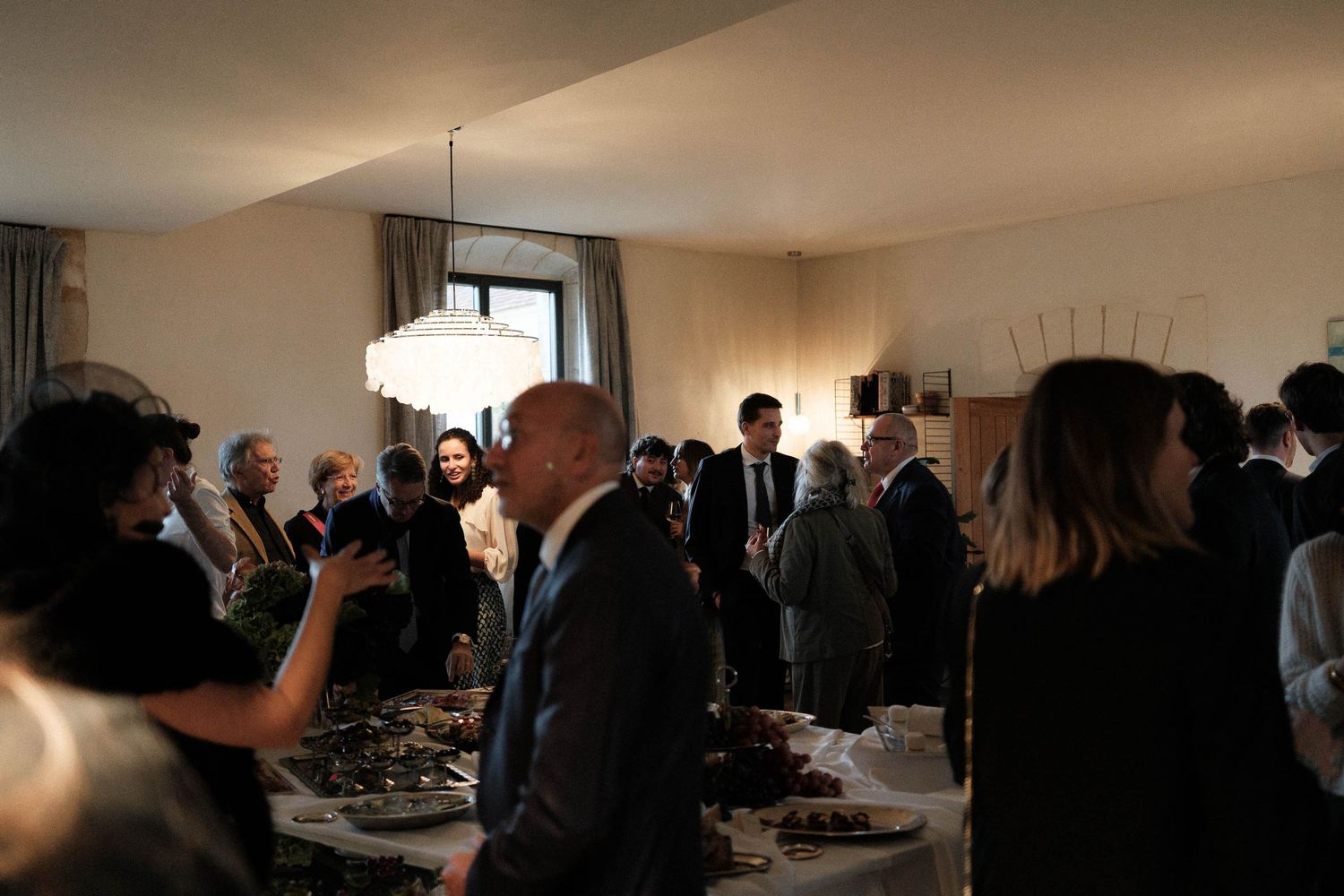 A group of people mingle and socialize at an indoor event with a buffet table and natural light coming through windows.
