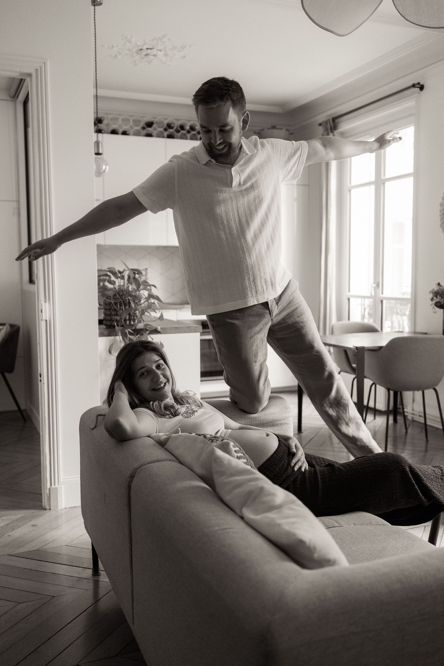 Two people play and interact playfully on a gray couch in a bright, minimalist living room setting.