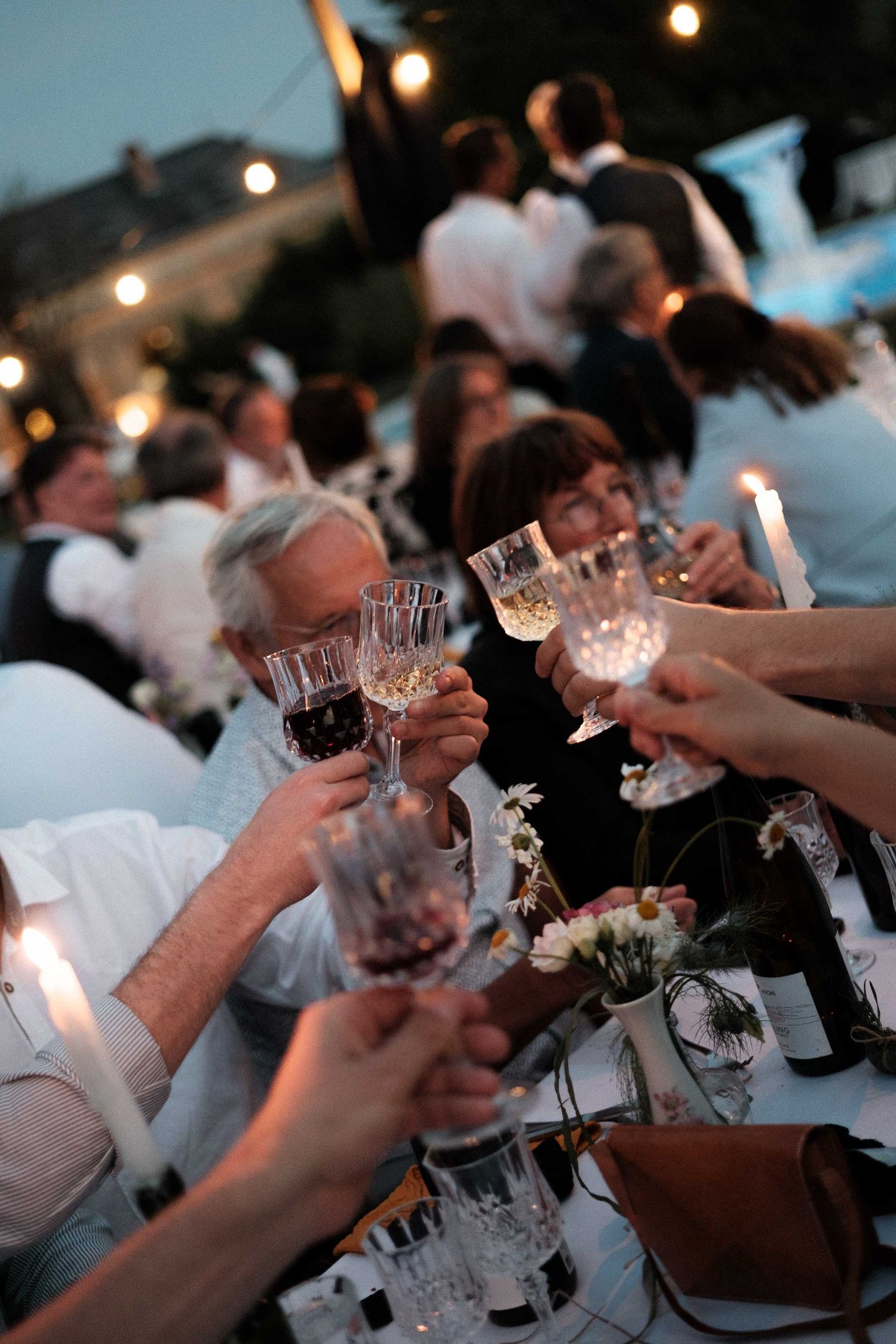 People raise wine glasses in a toast during an evening celebration with ambient lighting and white floral centerpieces.