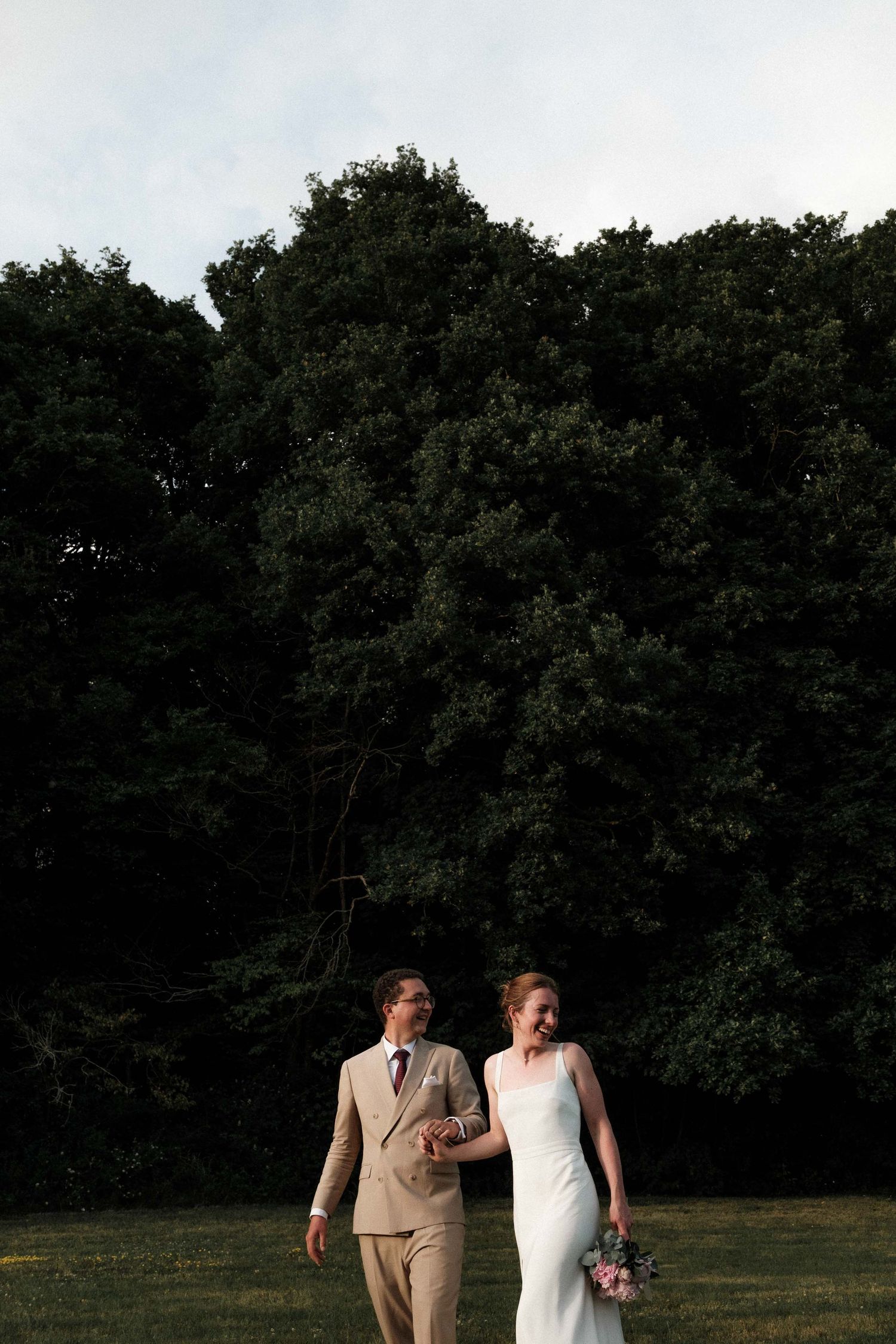A couple walks together through a grassy field with trees in the background during golden hour.
