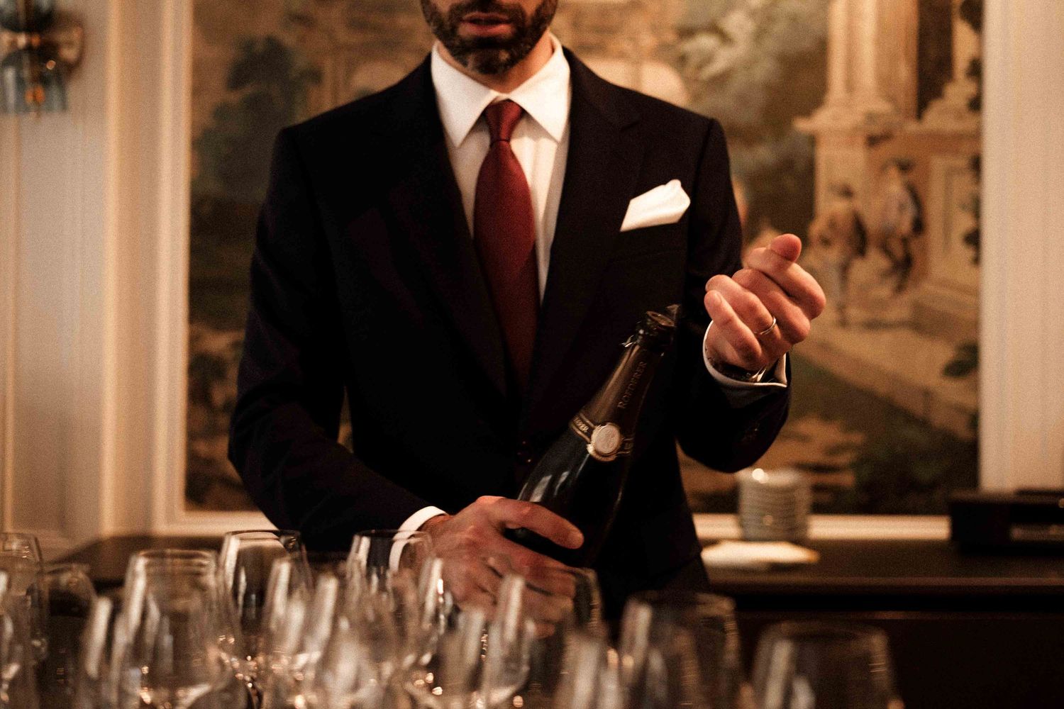 A person in a suit pours wine into glasses at an elegant bar setting with ornate decor in warm lighting.