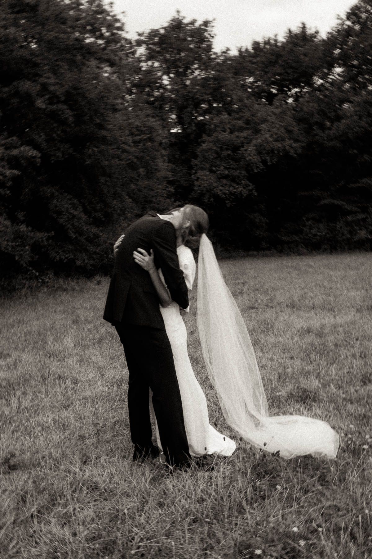 A romantic black and white wedding photograph shows a couple embracing in a meadow with trees in the background.