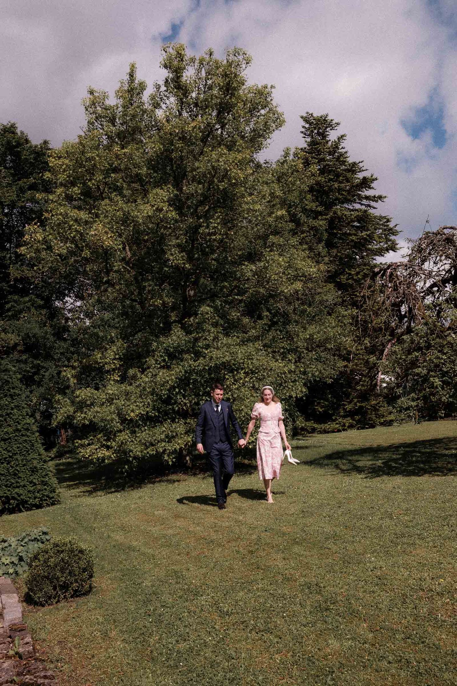 Two people walk together on a grassy hillside with trees in the background on a partly cloudy day.