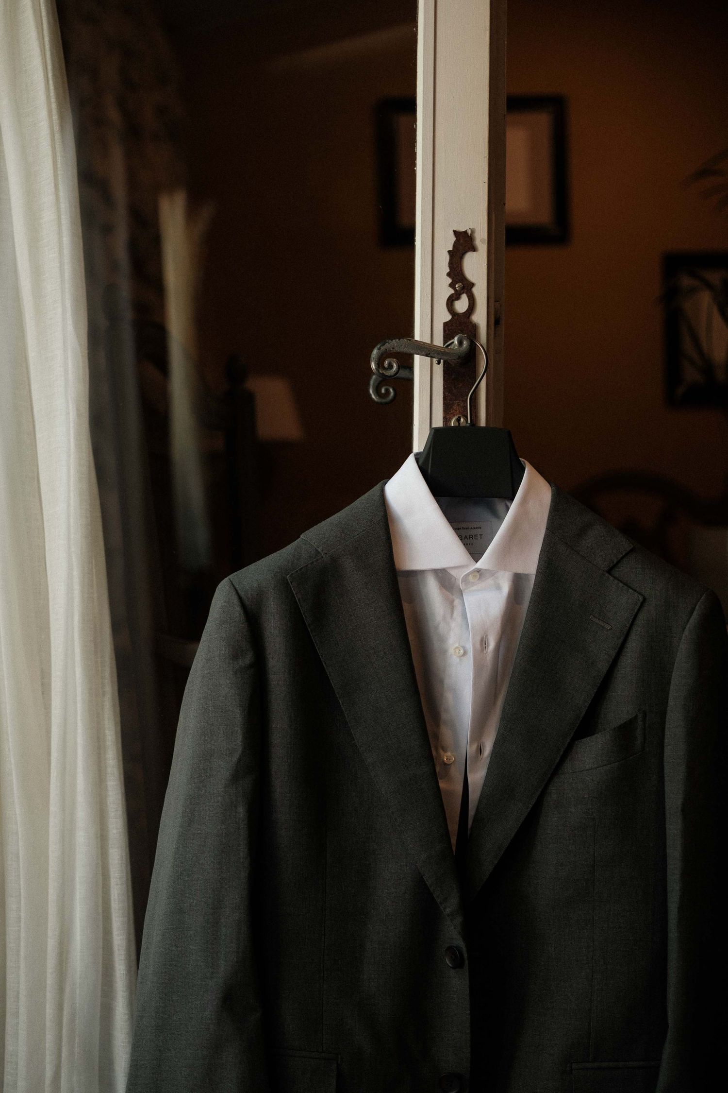 Multiple formal suit jackets arranged on dress forms near window with white curtains in moody lighting.