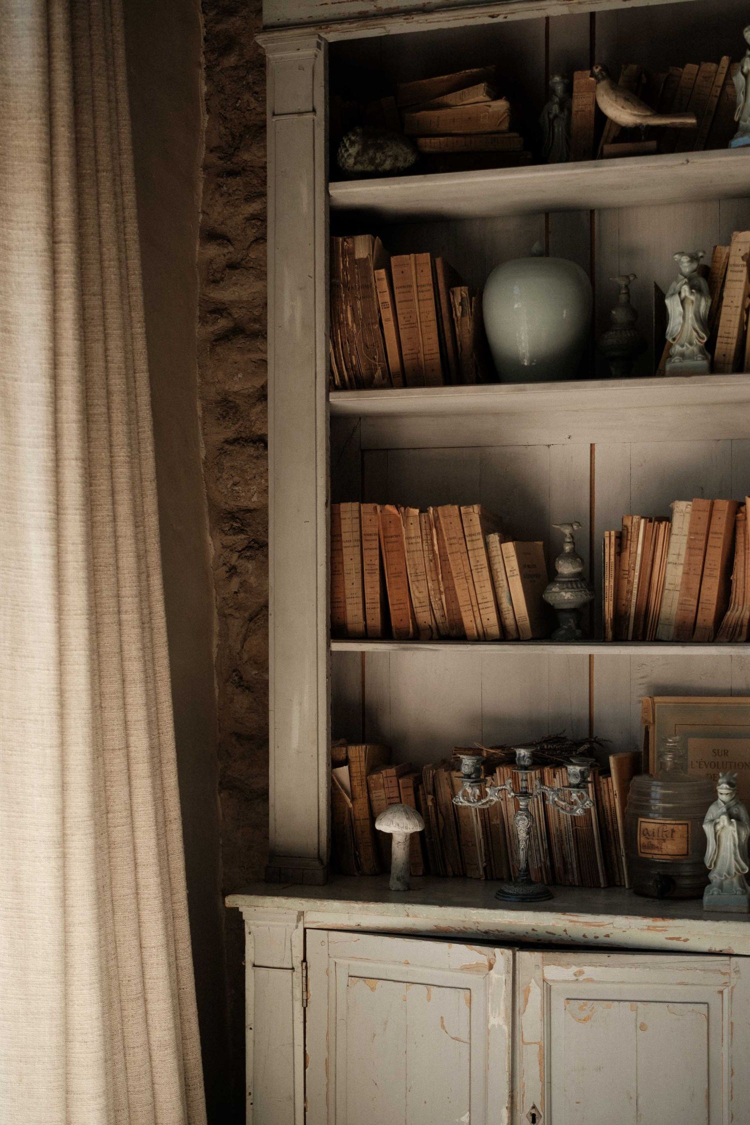 Built-in bookshelves with antique books and decorative objects against a neutral wall with flowing curtains.