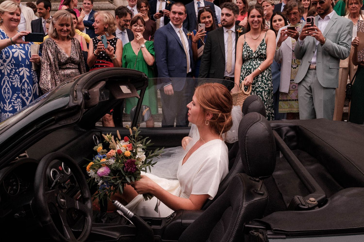 A bride in a white dress sits in a convertible car holding a bouquet while wedding guests look on and smile.