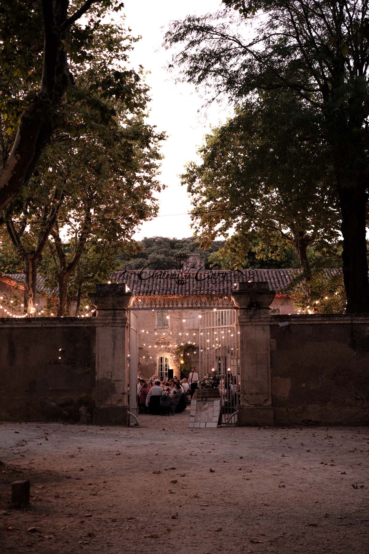 Stone entrance gate with string lights leading to an outdoor event venue at dusk with trees framing the scene.
