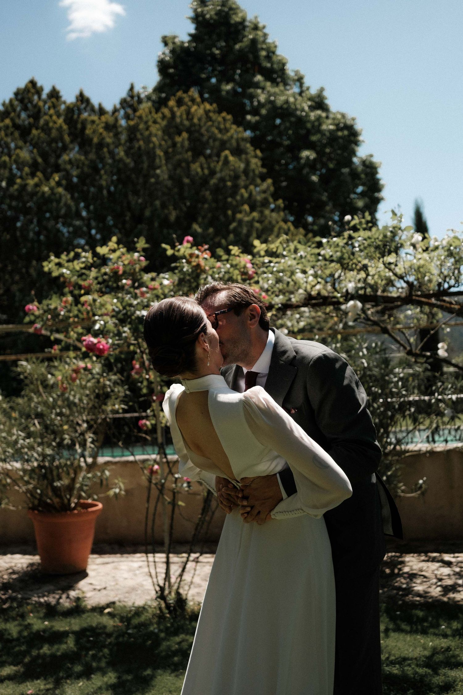A romantic wedding couple shares a tender moment in a garden with blooming roses and sunlit trees in the background.