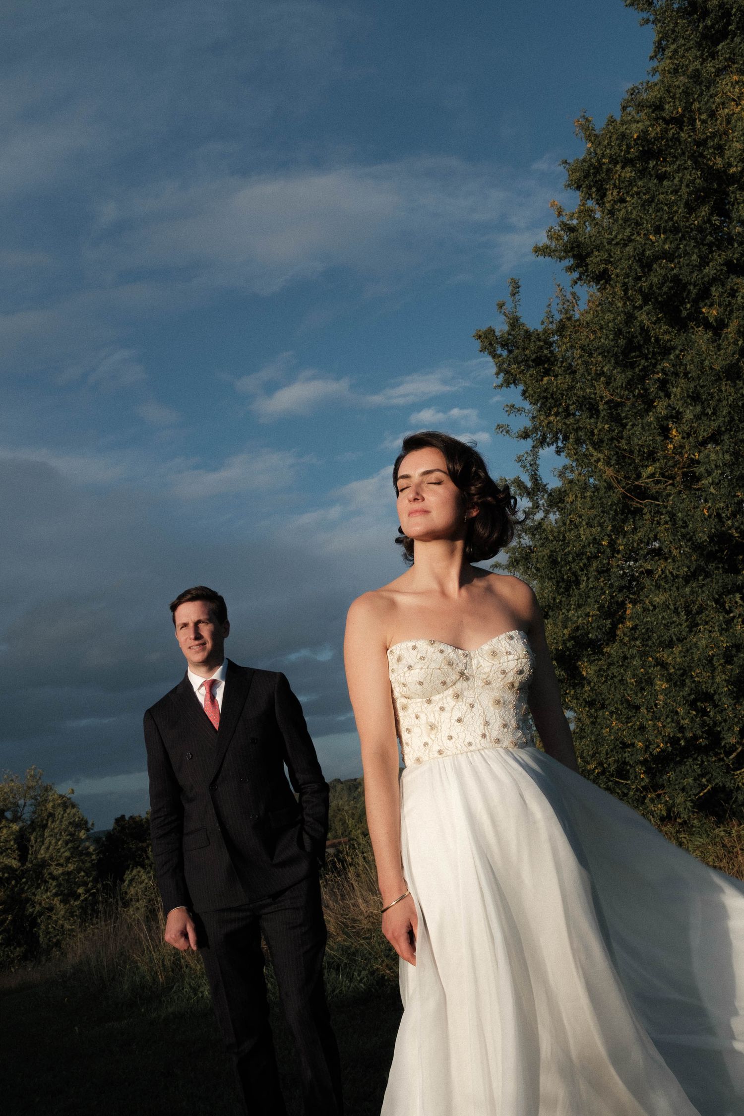 A bride in a white polka dot dress and groom in dark suit pose separately against a dramatic cloudy sky at sunset.