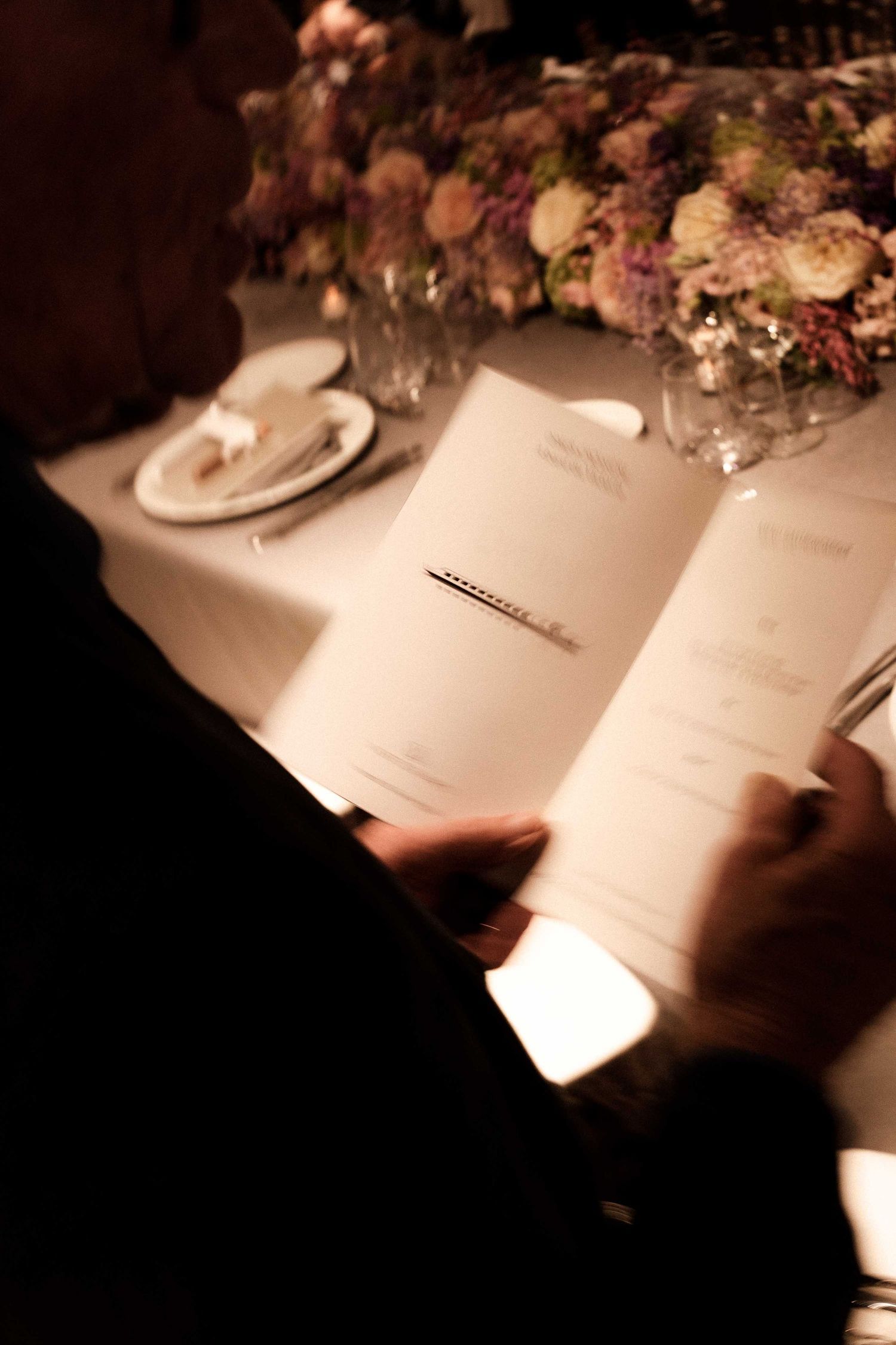 A person reads a menu at an elegant dining table with floral centerpiece and place settings in warm lighting.
