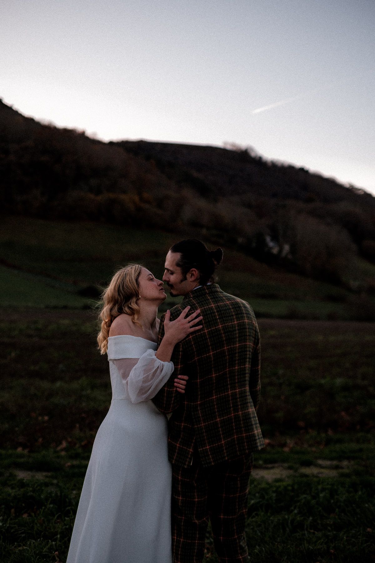 A romantic couple embraces and kisses in a scenic outdoor field at dusk with rolling hills in the background.