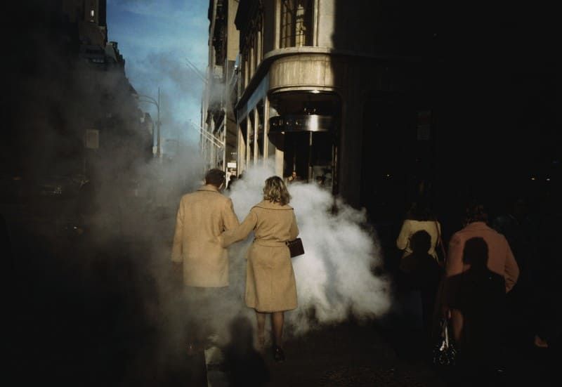 Silhouettes of people walking through atmospheric fog and steam on a dark city street at night.