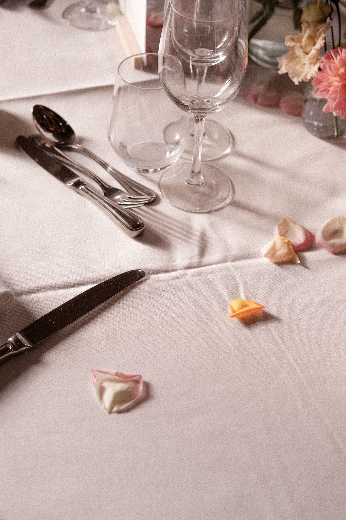 Elegant table setting with wine glasses, silverware, and scattered flower petals on white linen tablecloth.