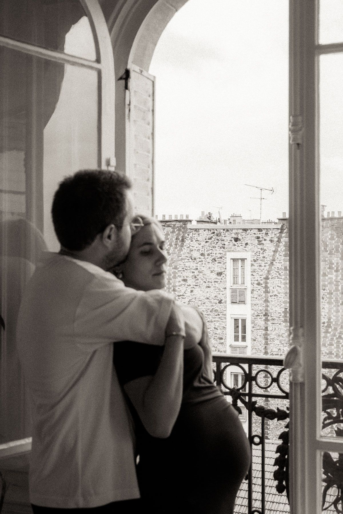 A romantic black and white photograph of a couple embracing on a Parisian balcony overlooking stone buildings.