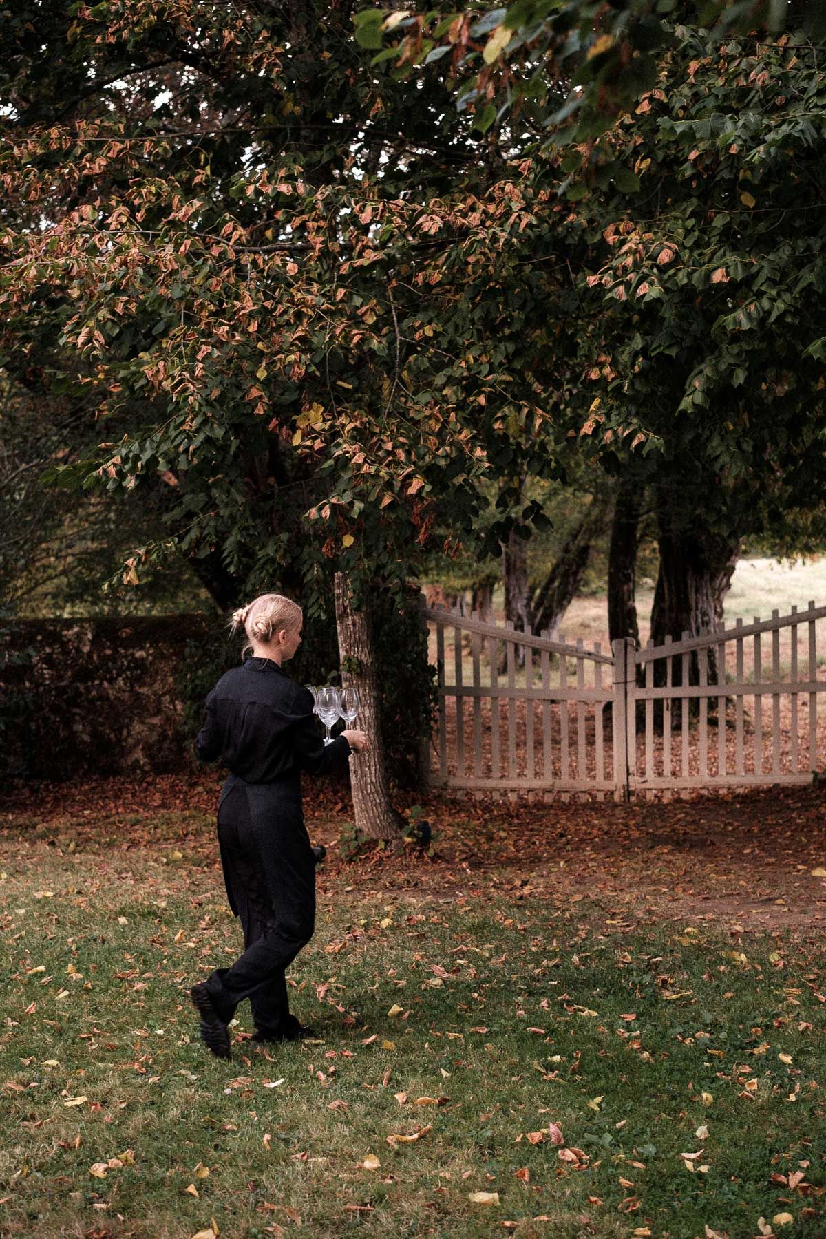 A person in dark clothing walks through an autumn yard with fallen leaves and a wooden fence in the background.