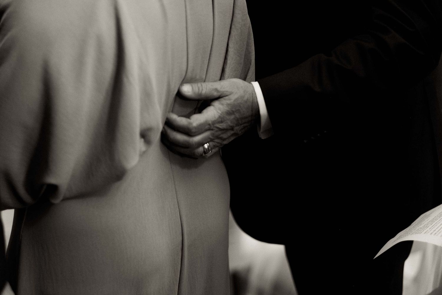A close-up black and white photograph shows hands clasped behind someone's back wearing formal attire.
