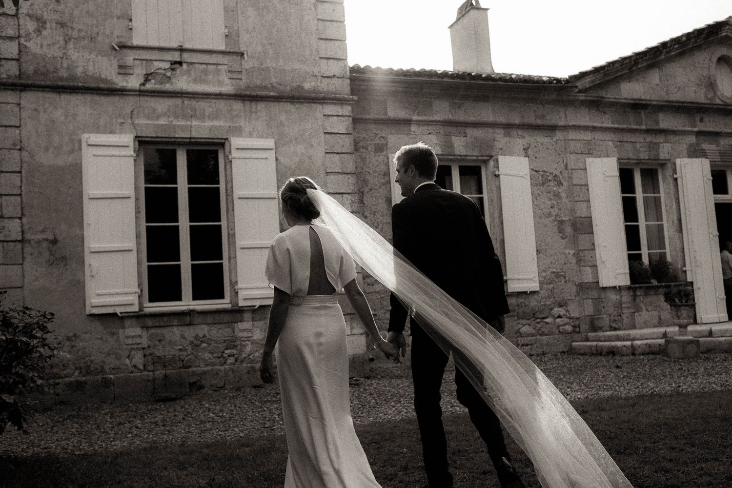 A romantic black and white wedding photo showing a flowing veil trailing behind a couple outside a stone building.