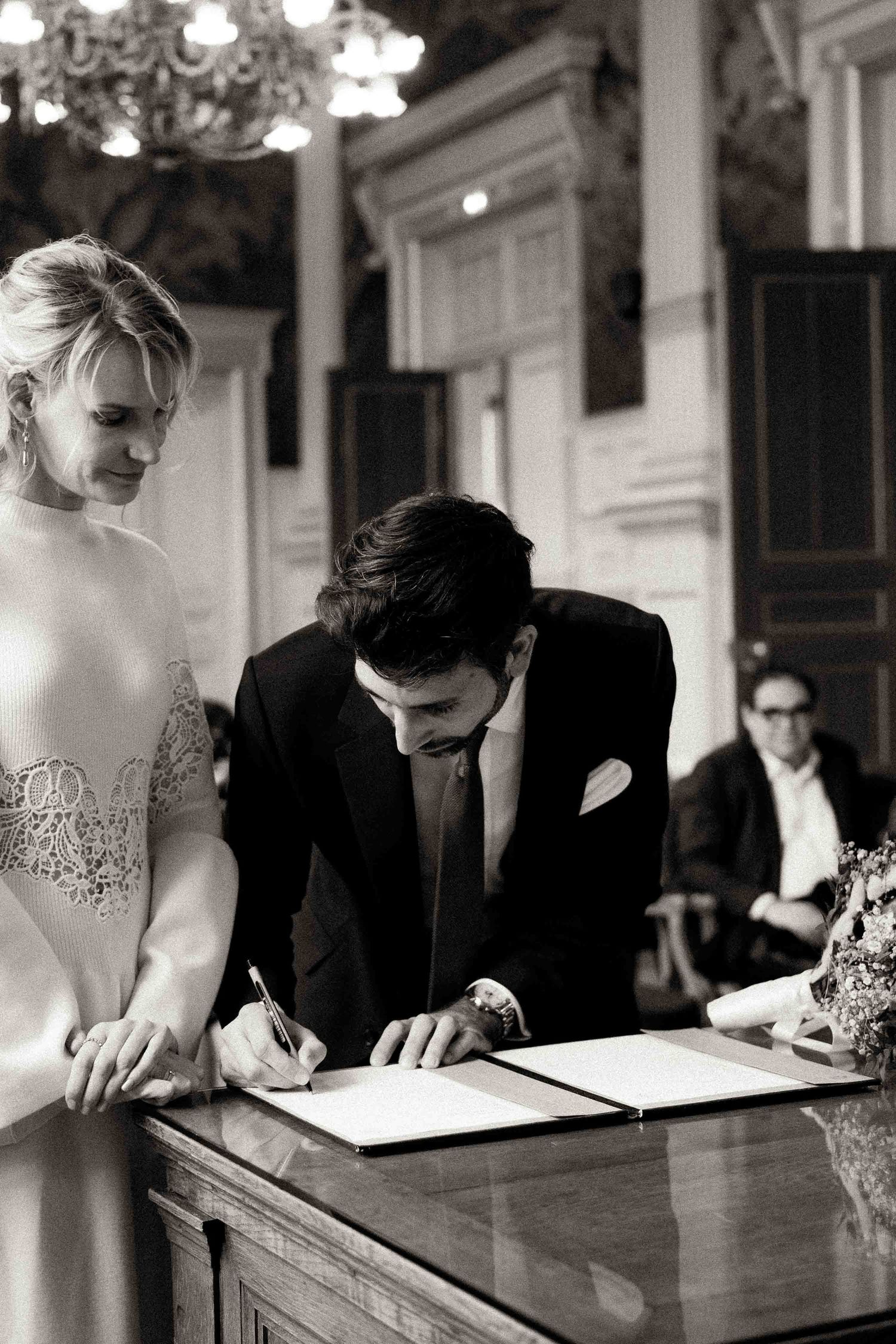 A black and white photo shows people signing documents at an elegant venue with chandeliers overhead.