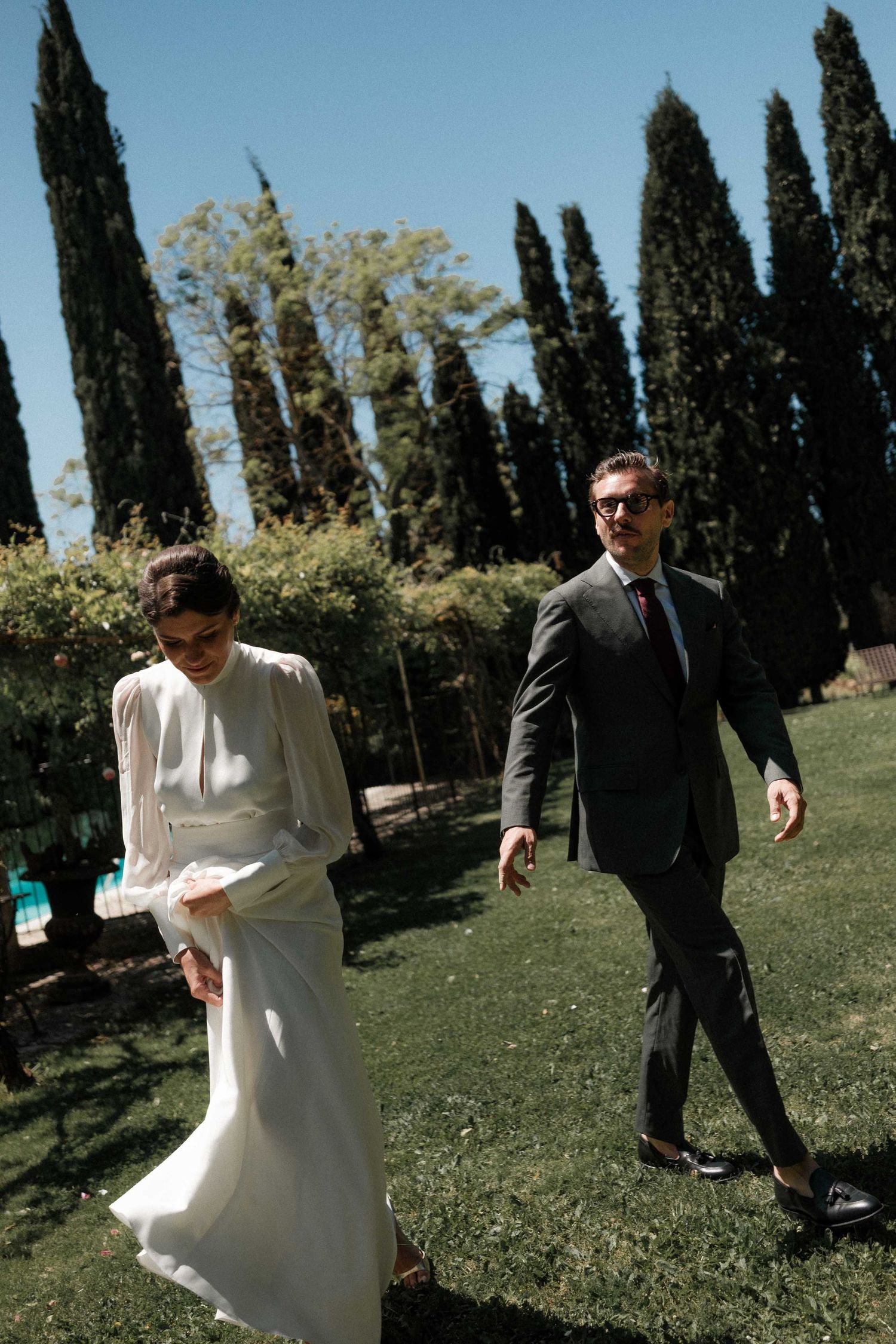 A couple in wedding attire walking through a garden path lined with tall cypress trees.