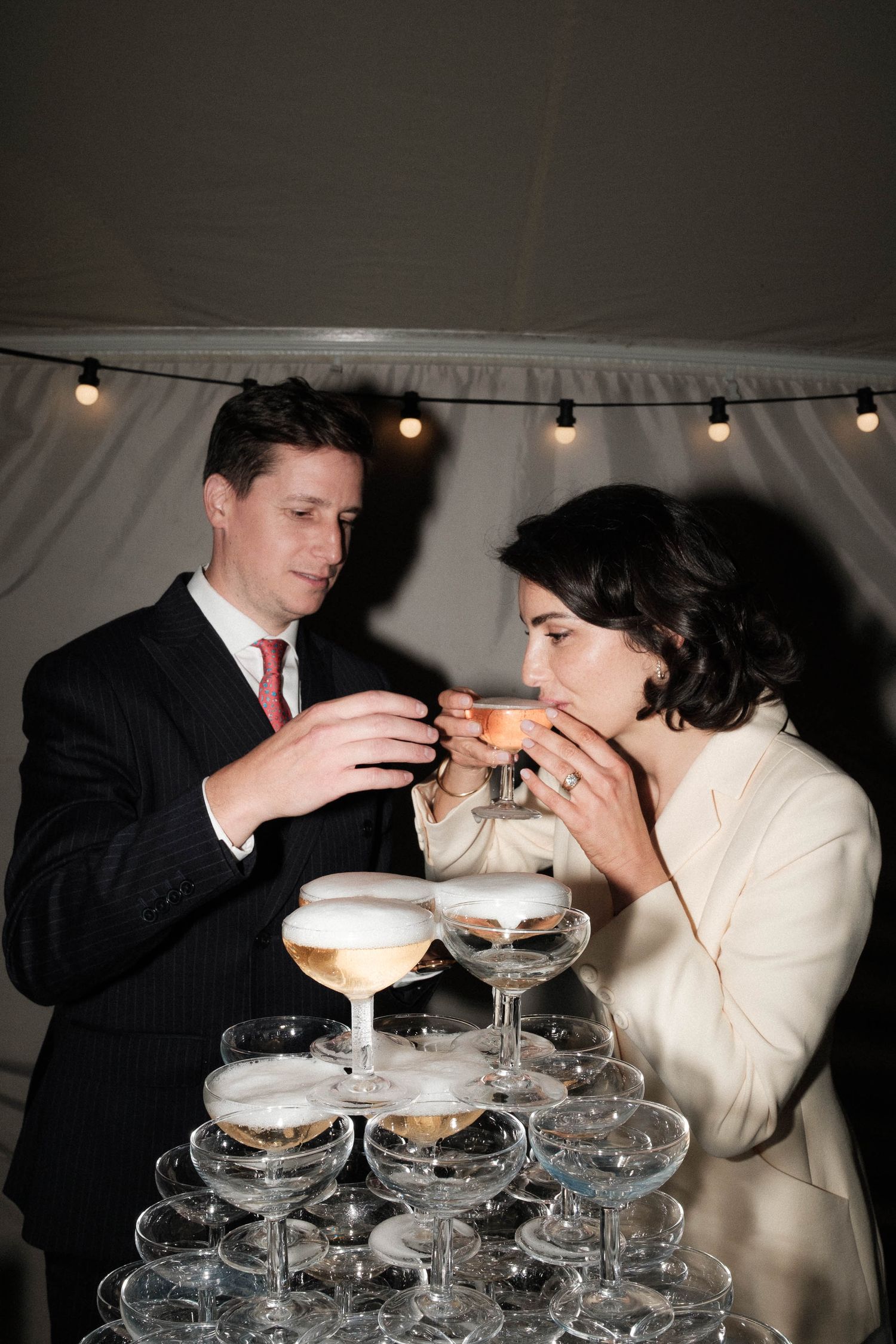A couple pours champagne into a tower of stacked coupe glasses at an elegant celebration.
