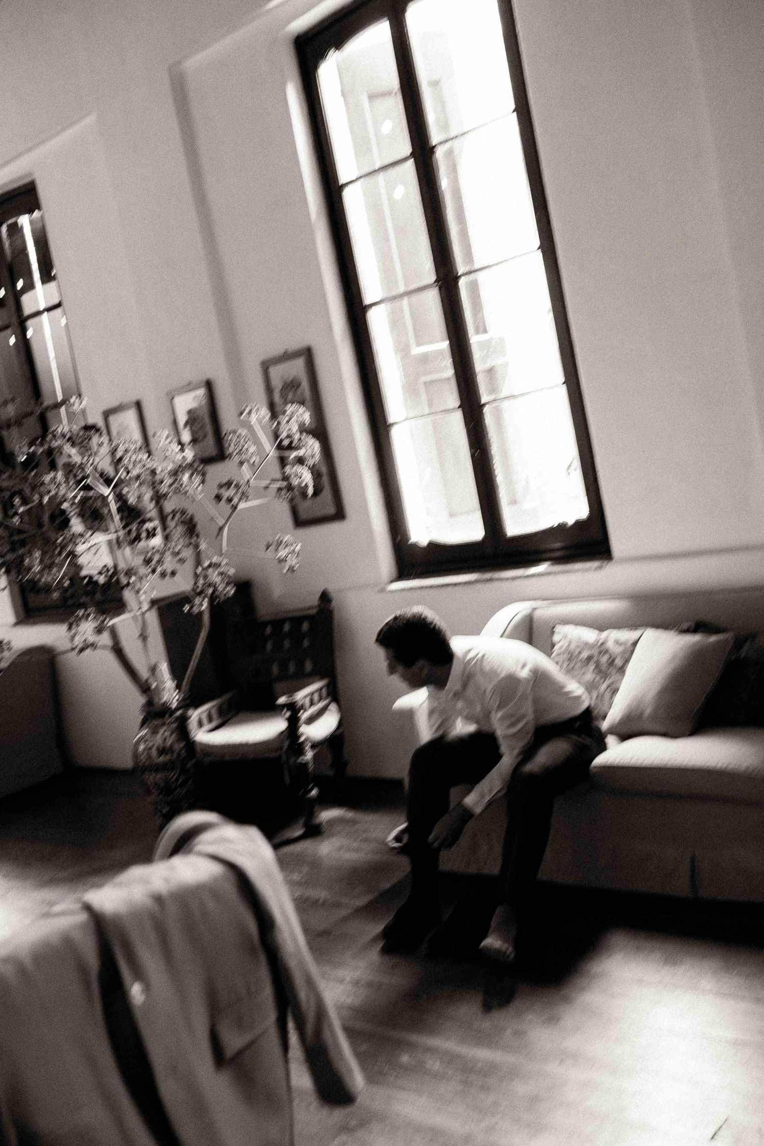 Black and white interior photo showing a figure sitting on a couch near a tall window in a sparsely decorated room.