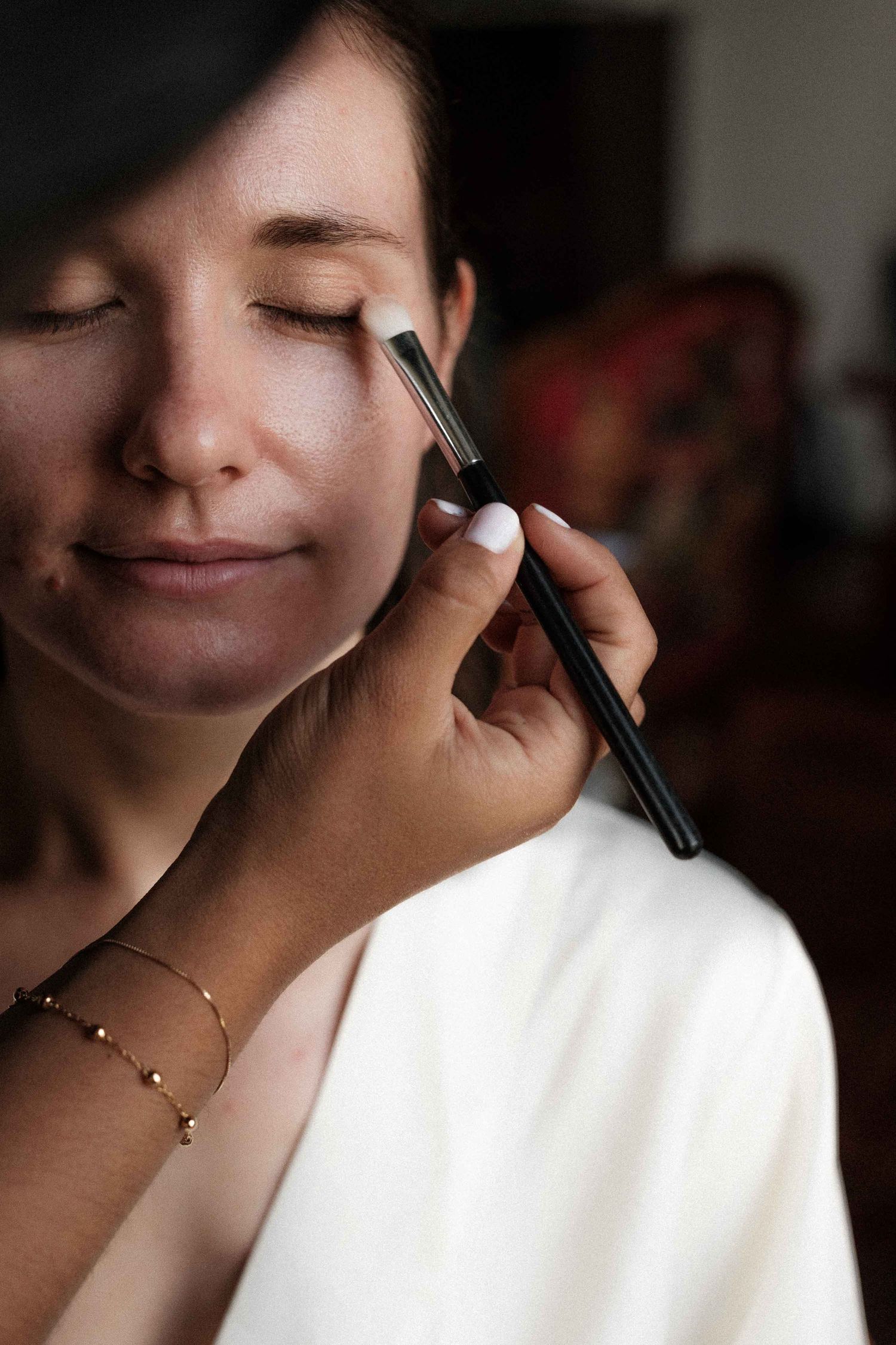 Makeup artist applies eyeshadow with a brush while client relaxes with eyes closed in white robe.