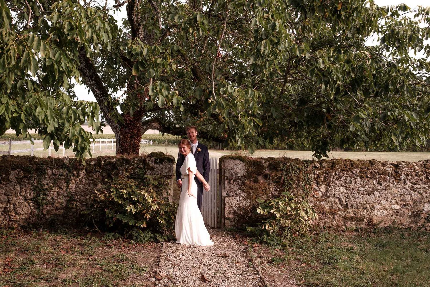 A couple shares an intimate moment against a rustic stone wall and tree in a romantic garden setting.