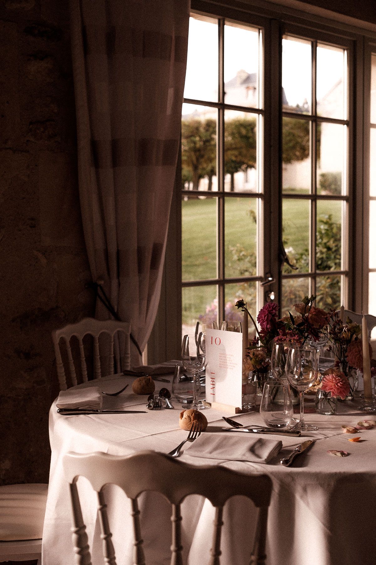 Elegant table setting with white linens, wine glasses and candles near a window overlooking pastoral grounds at dusk.
