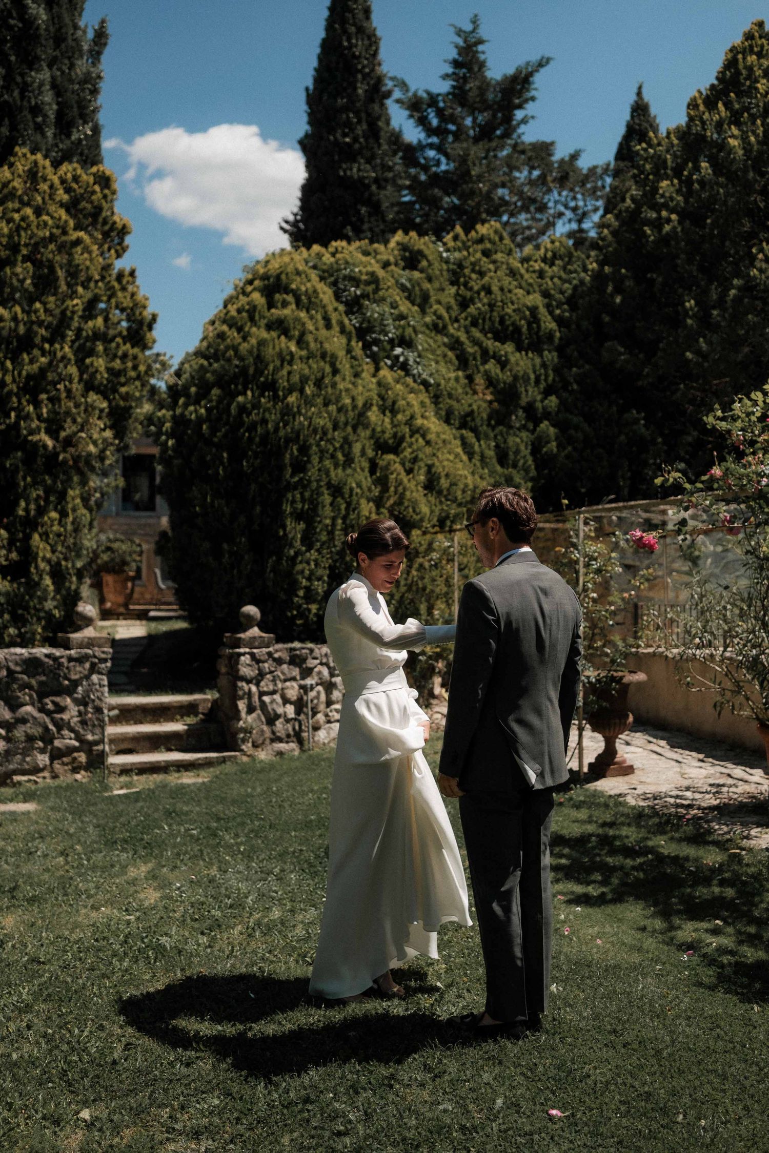 A couple shares an intimate moment in a garden setting with cypress trees and stone walls.