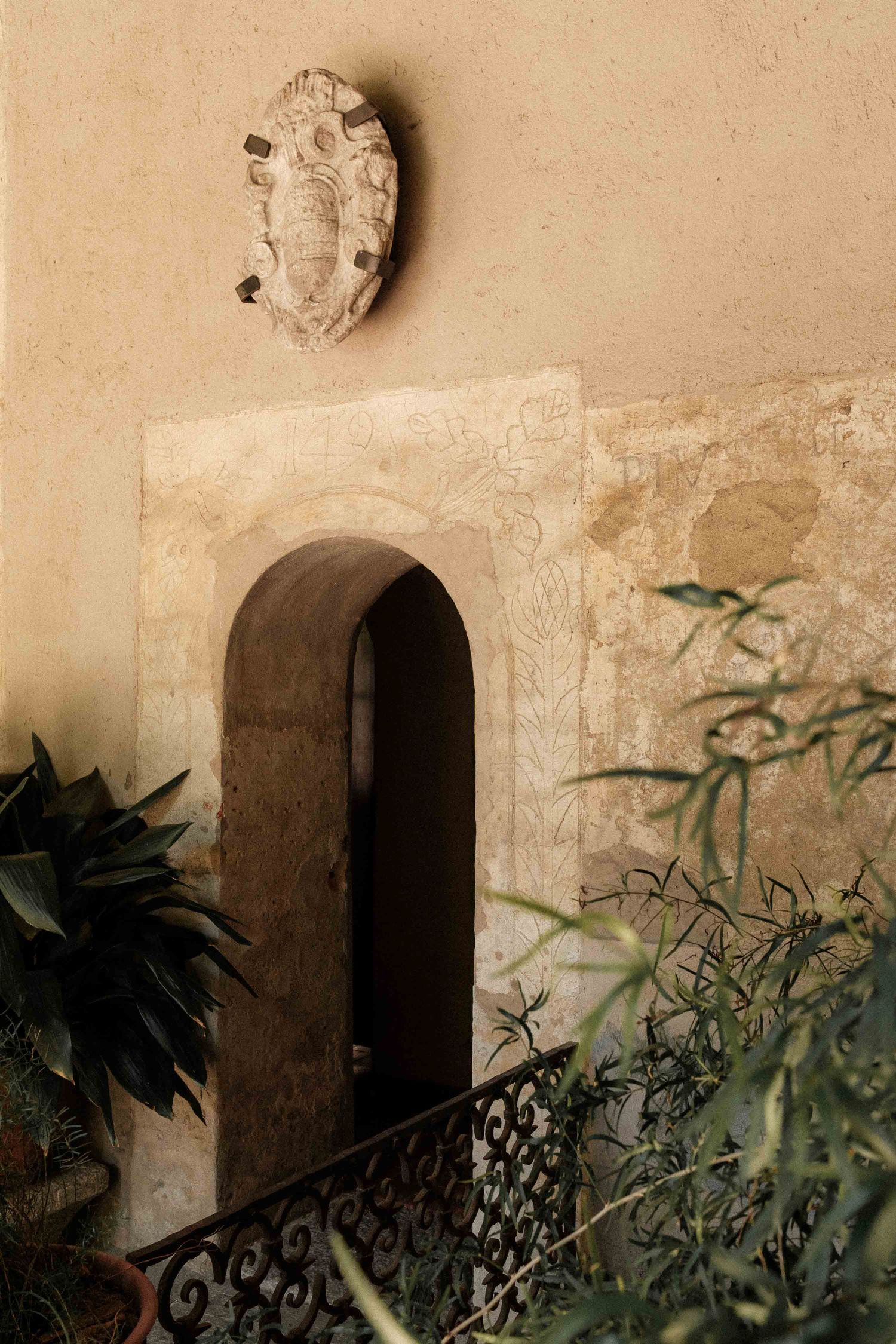 Ancient stone wall with arched doorway and decorative wrought iron details partially obscured by green foliage.