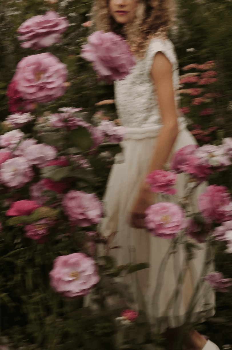 A person in a white dress stands among blooming pink roses in a garden setting.