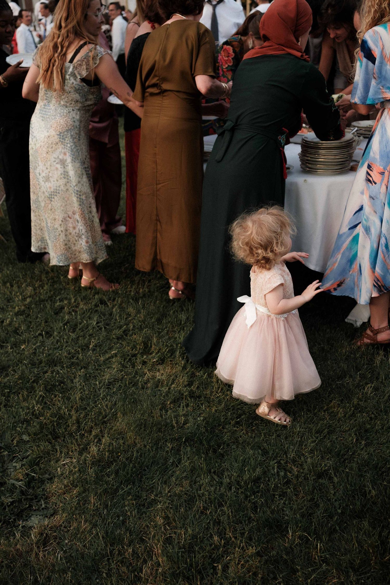 A small child in a pink tulle dress walks on grass during an outdoor gathering of people.