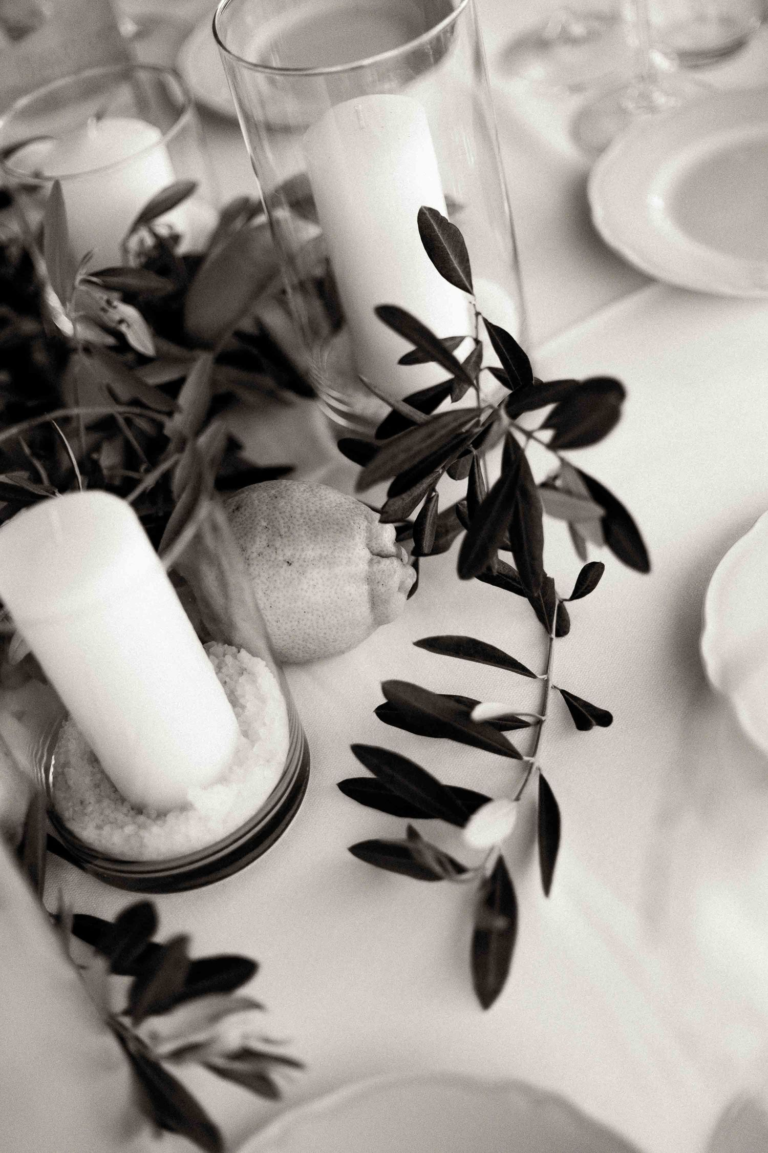 Black and white detail of wedding table decor featuring white candles and olive branch centerpiece on white tablecloth.