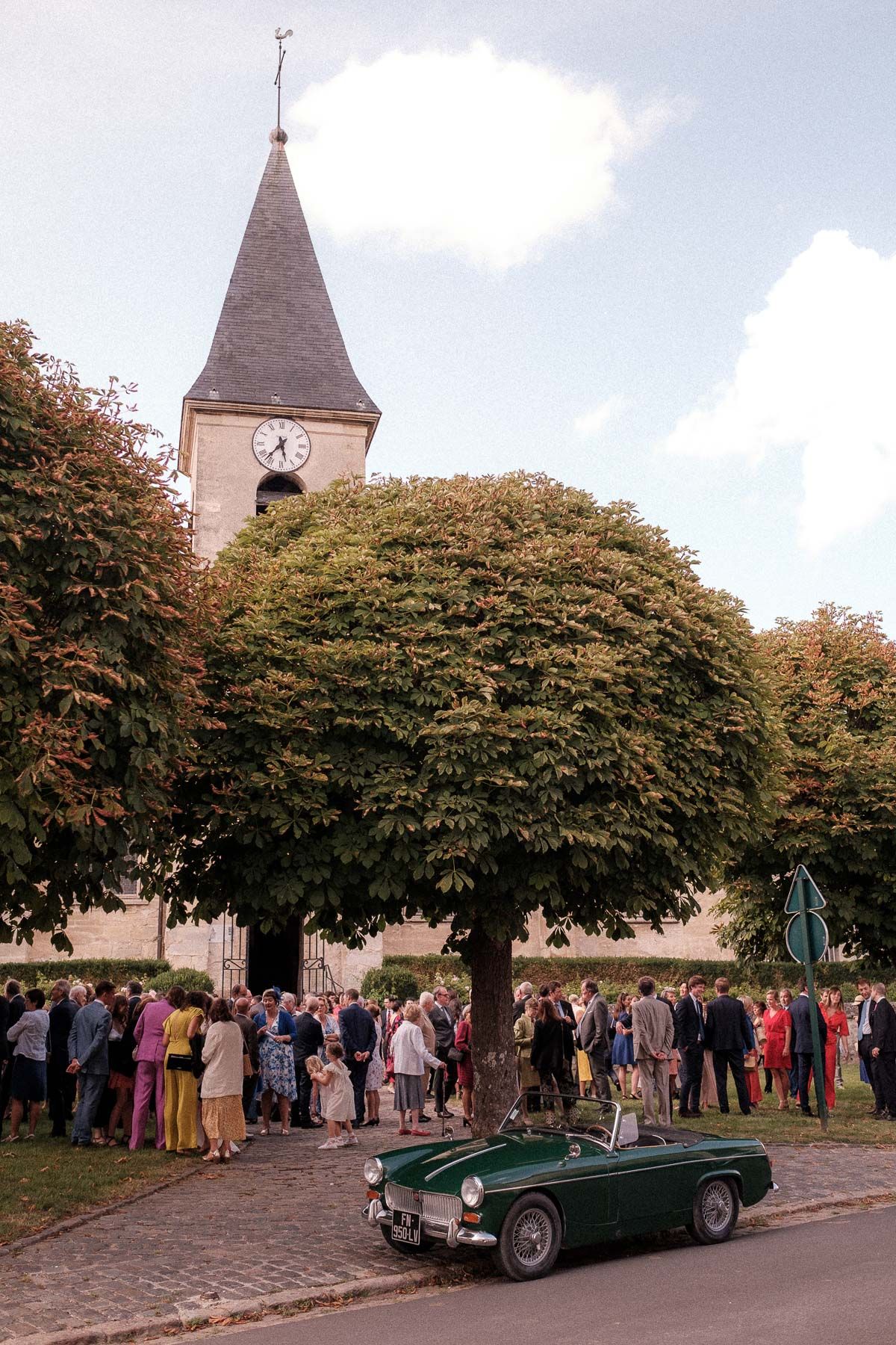 Vintage green MG sports car parked outside church with crowd gathered under large trees during autumn event.