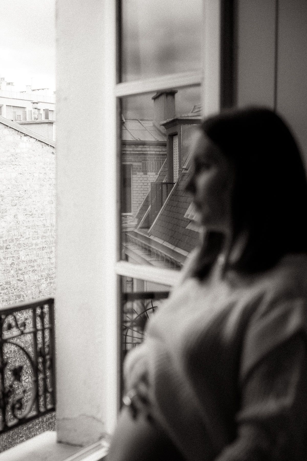 A silhouetted figure gazes pensively through a window at urban rooftops and fire escapes in black and white.