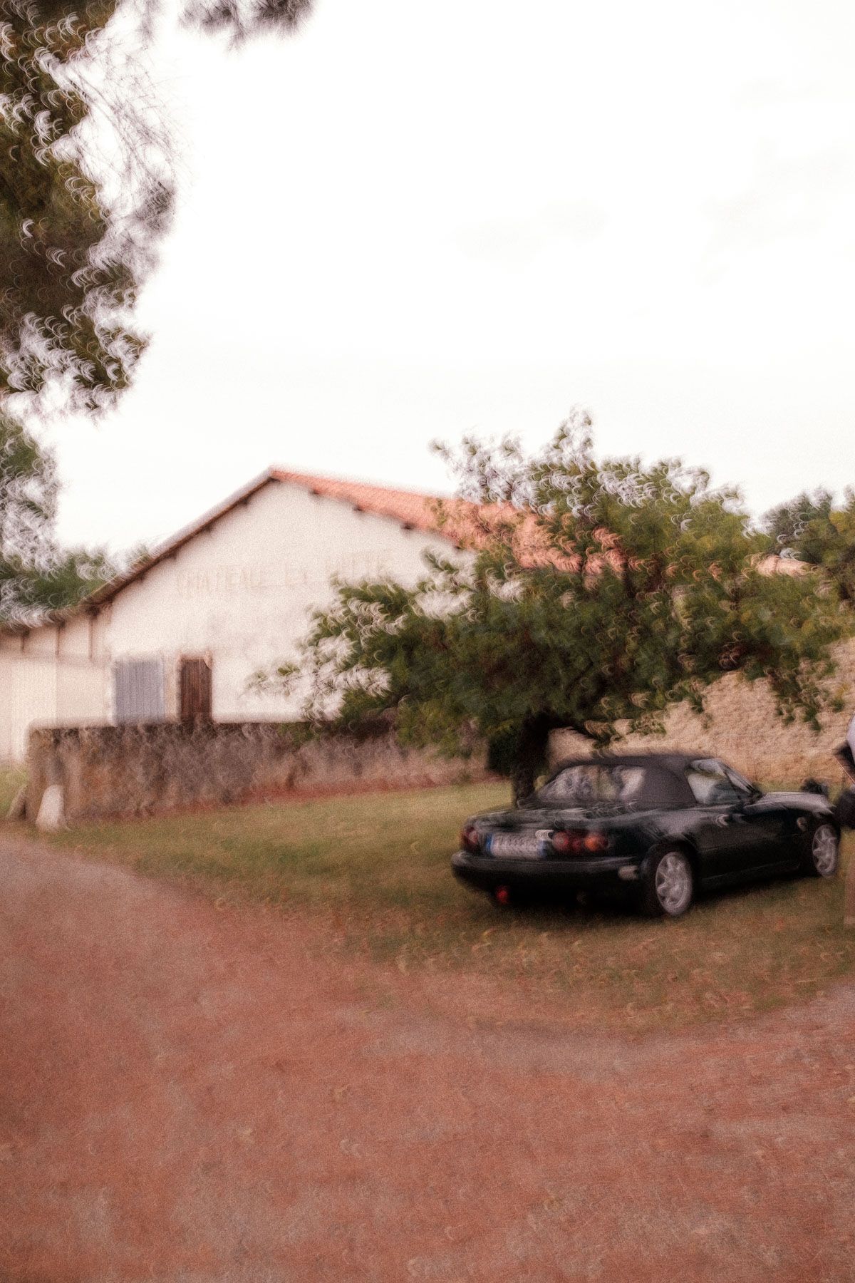 A black sedan car parked on dirt ground near a white building with trees in the background.