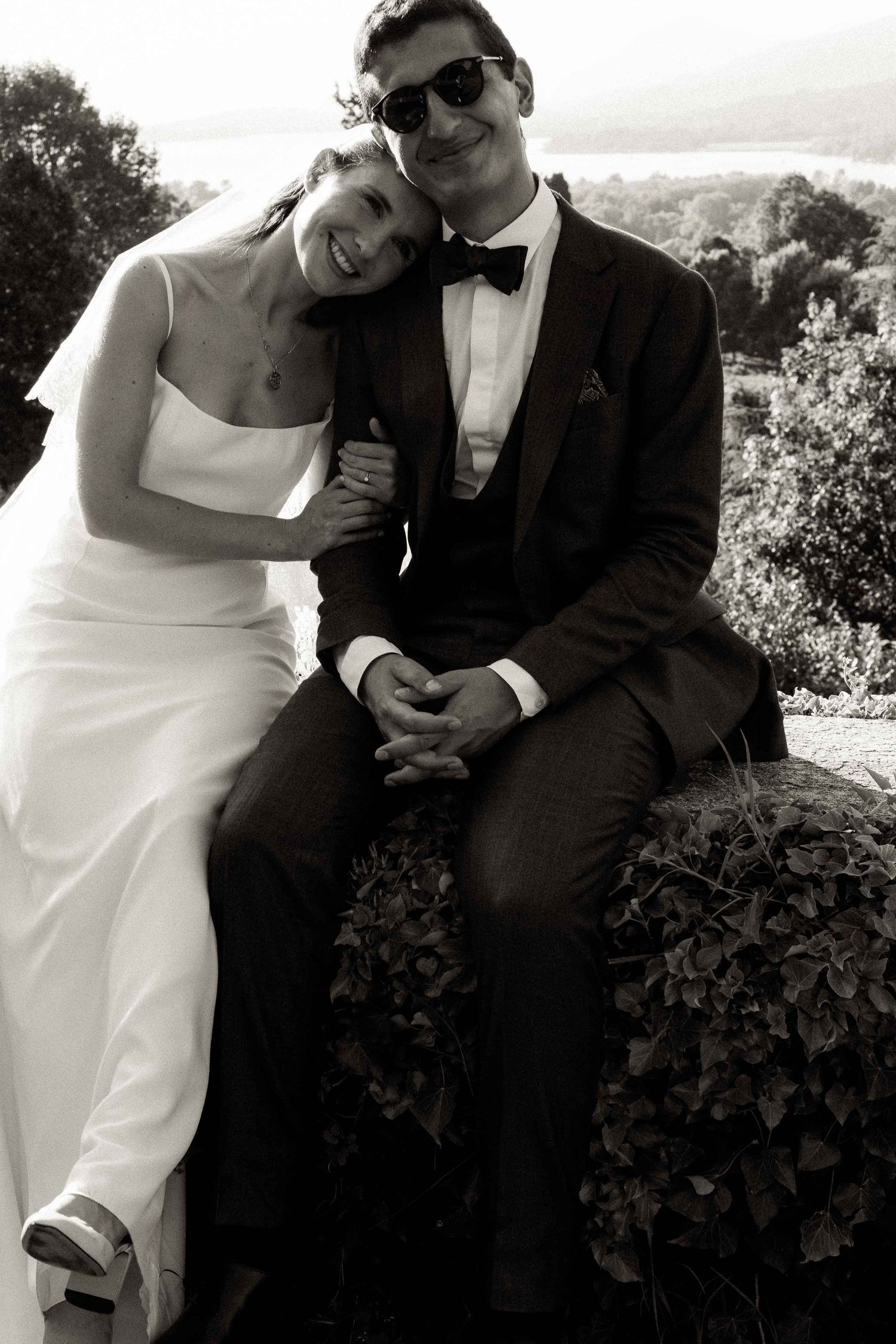 A black and white wedding photo series showing a couple in formal attire posing playfully outdoors against a scenic backdrop.