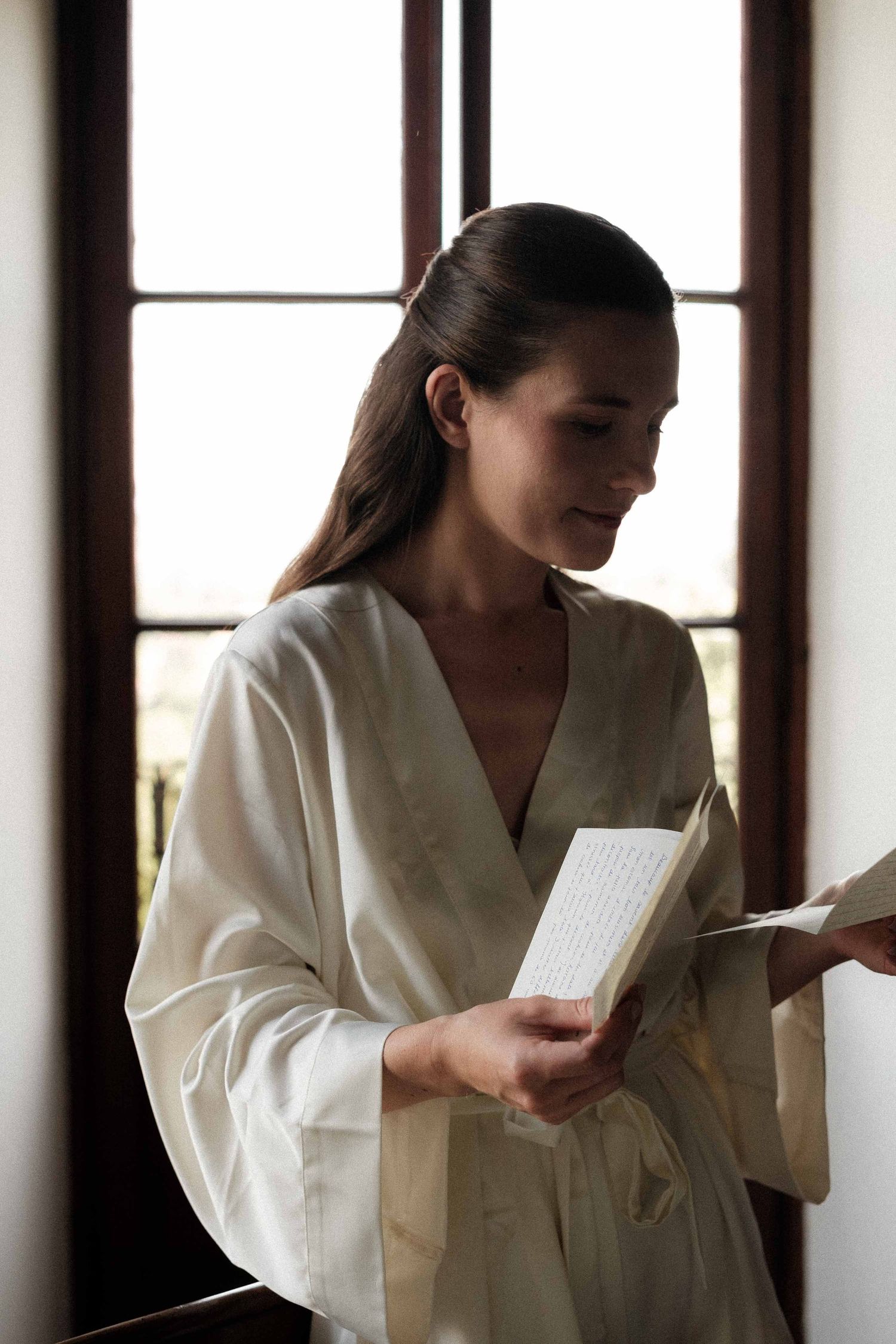 A person in a white bathrobe reads a book while standing near windows in a serene, sunlit room.